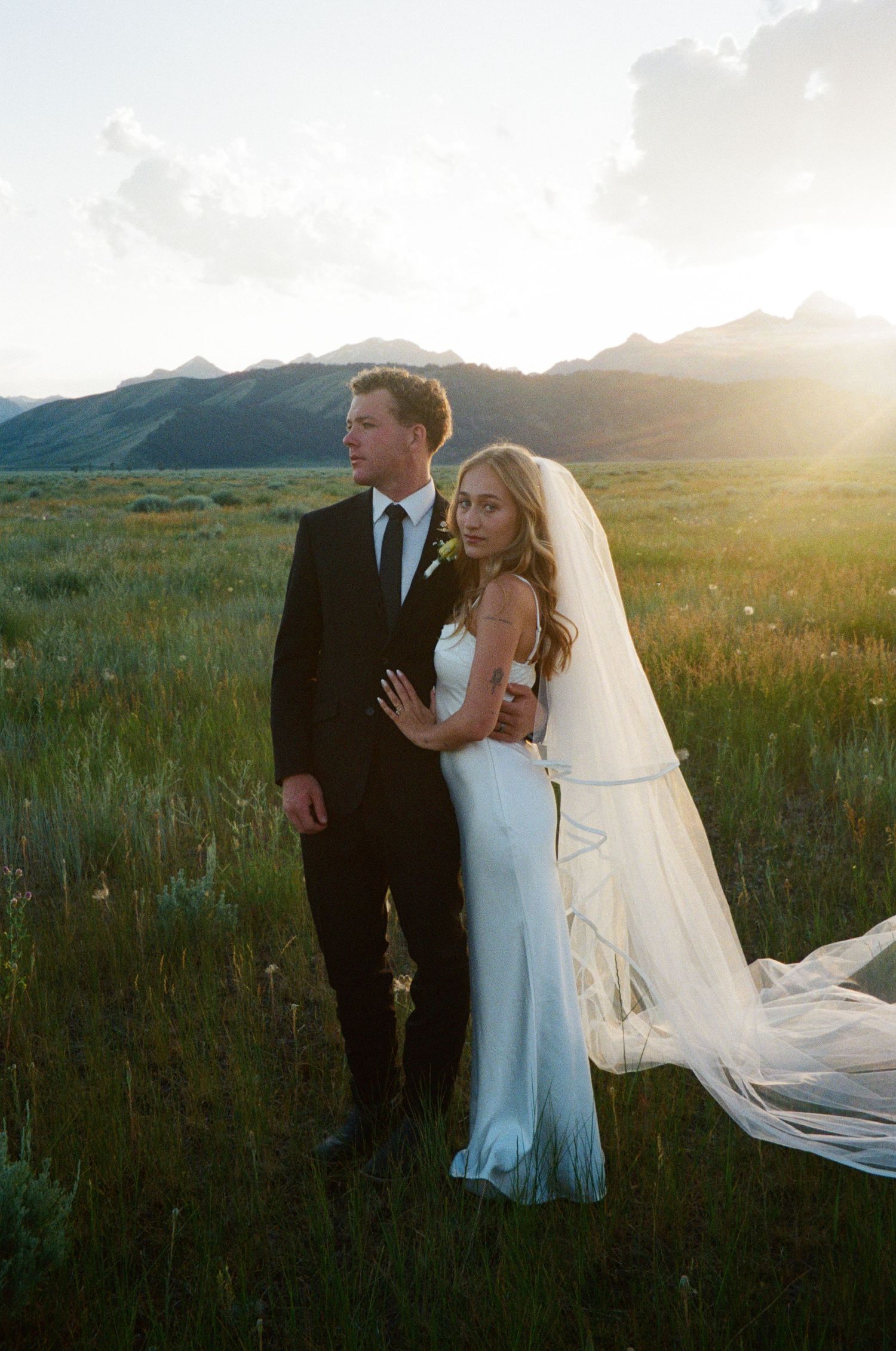 Bride and groom embrace in golden sunset meadow with dramatic mountain range backdrop.