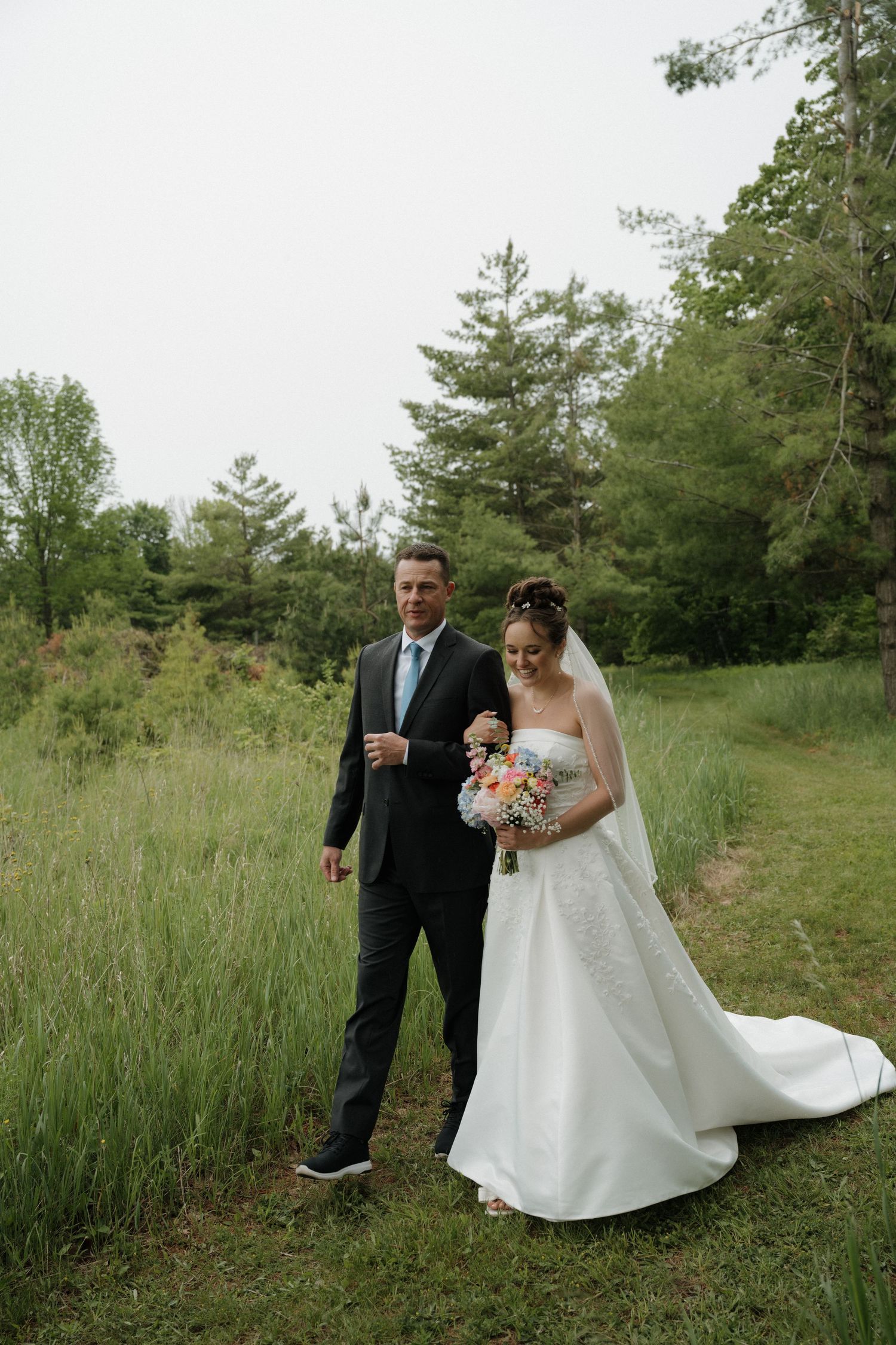 Bride in white gown walks with man in suit through lush greenery.