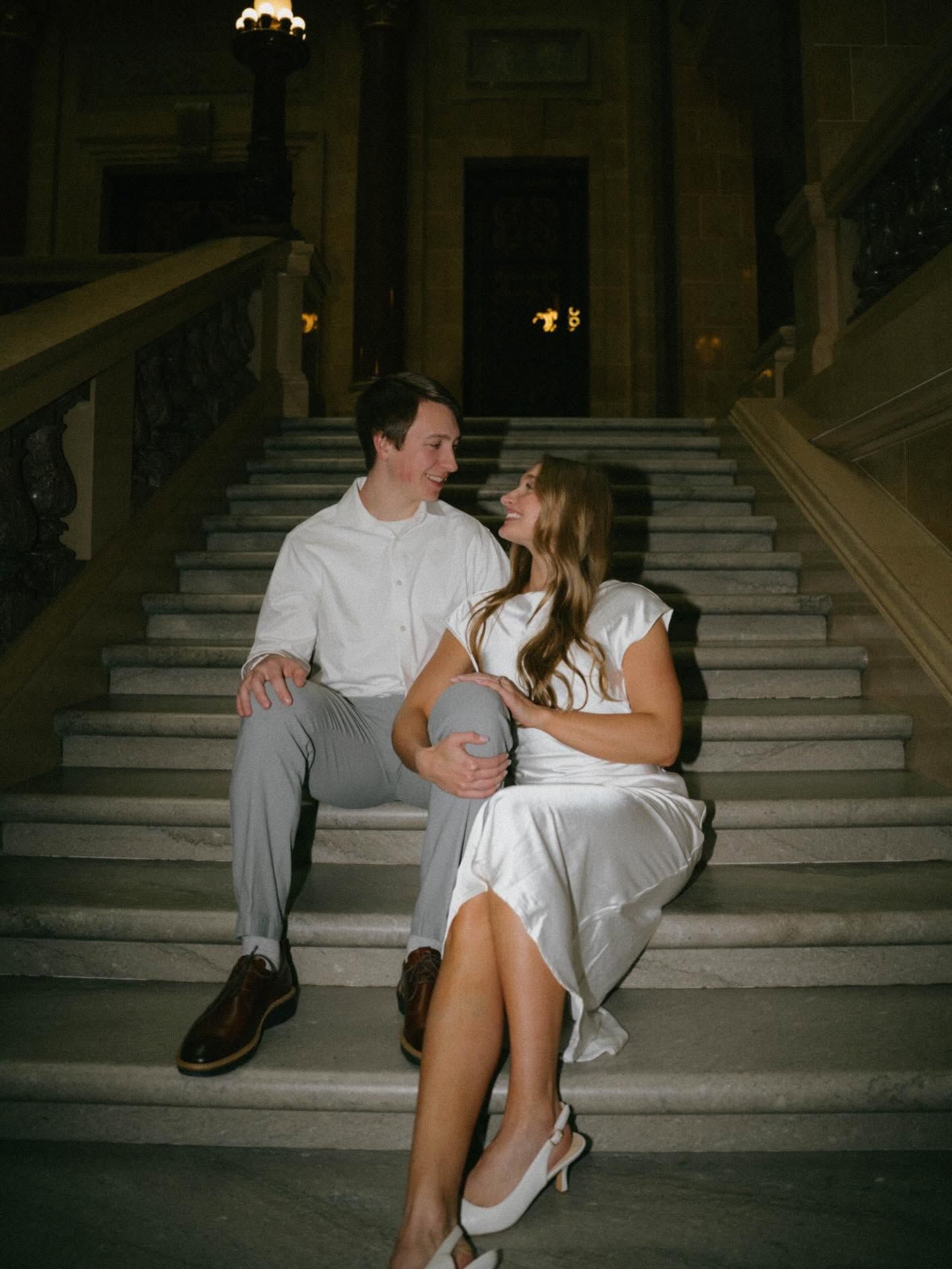 Couple sitting on marble stairs in elegant building, smiling at each other, woman in white dress, man in white shirt.