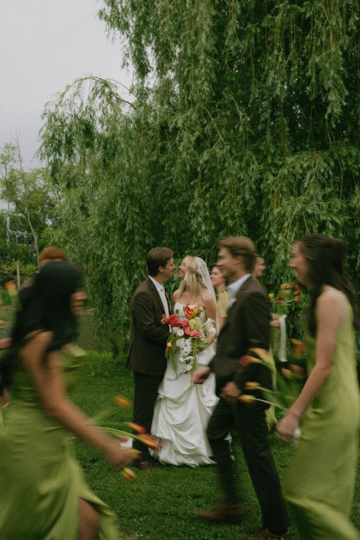 Bride and groom stand under a weeping willow tree surrounded by wedding party in green dresses during outdoor ceremony.
