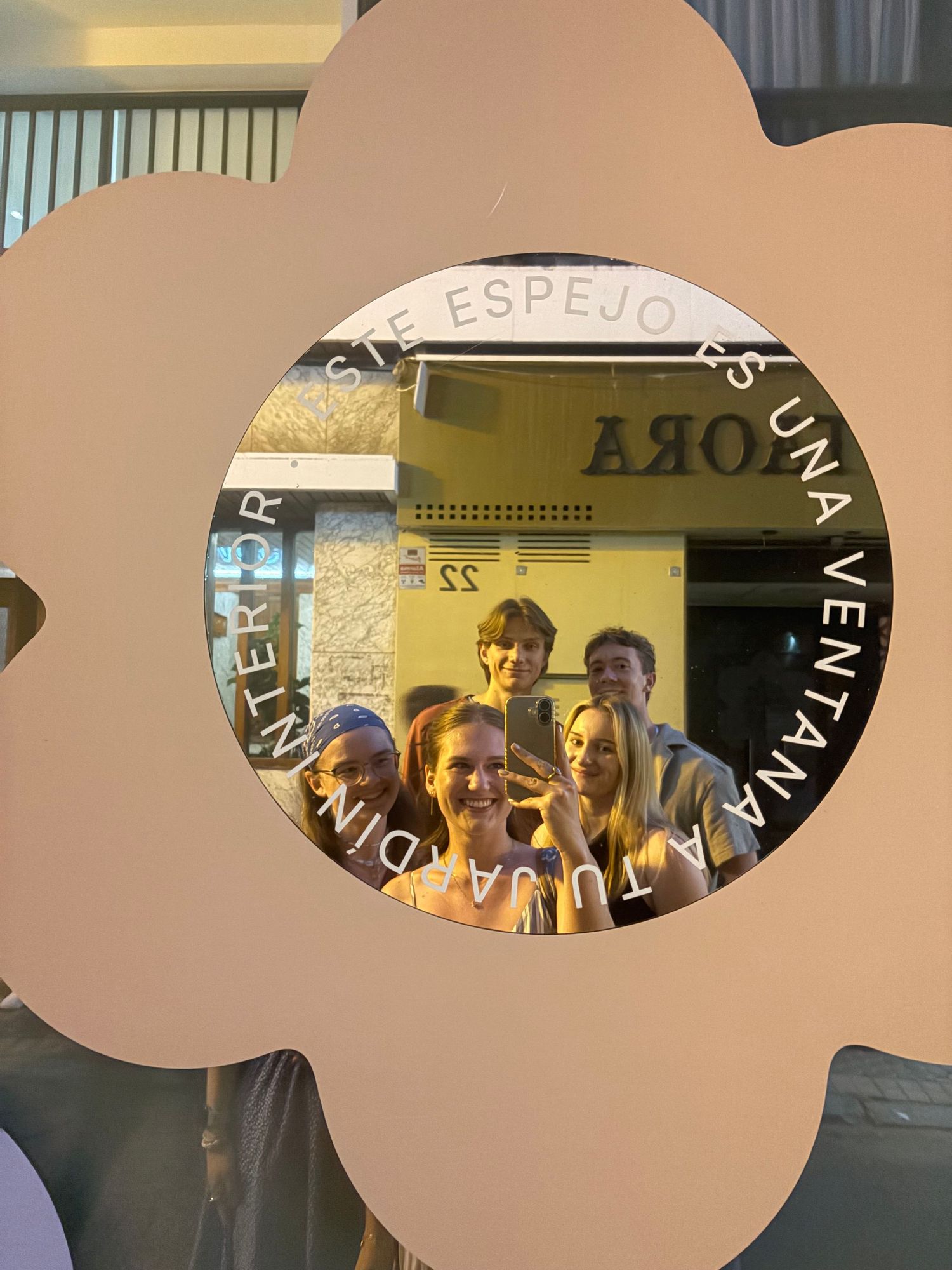 Group of friends taking selfie in flower-shaped mirror with Spanish text at trendy retail store.