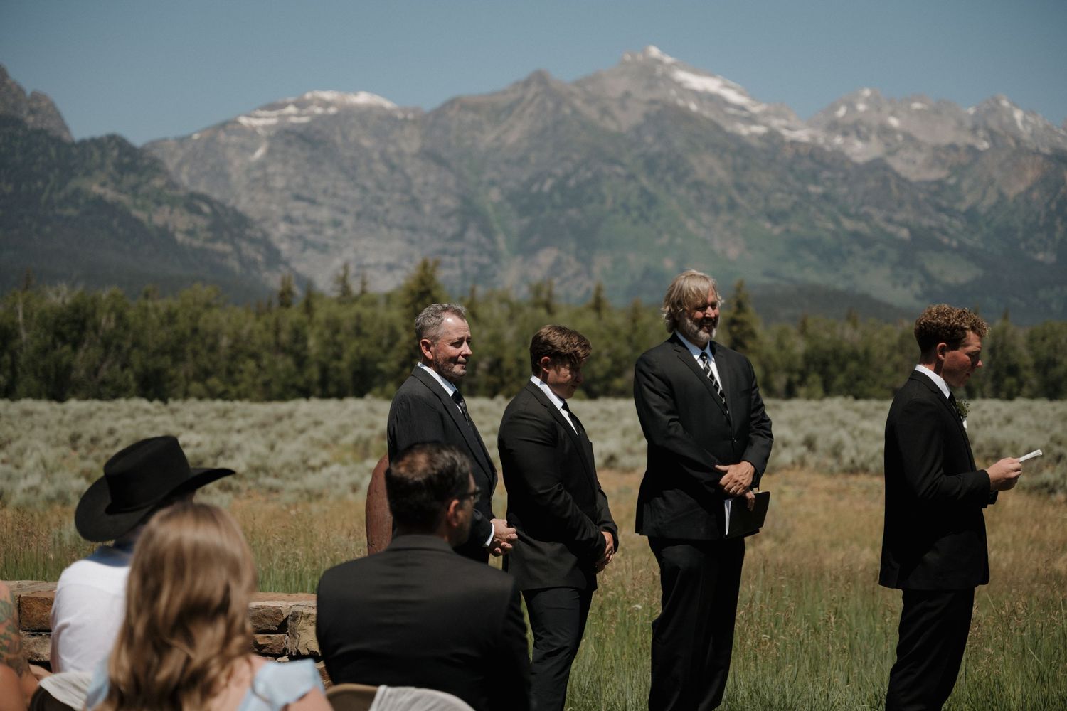 Groomsmen in black suits standing and chatting before outdoor wedding ceremony with Grand Teton mountains behind.