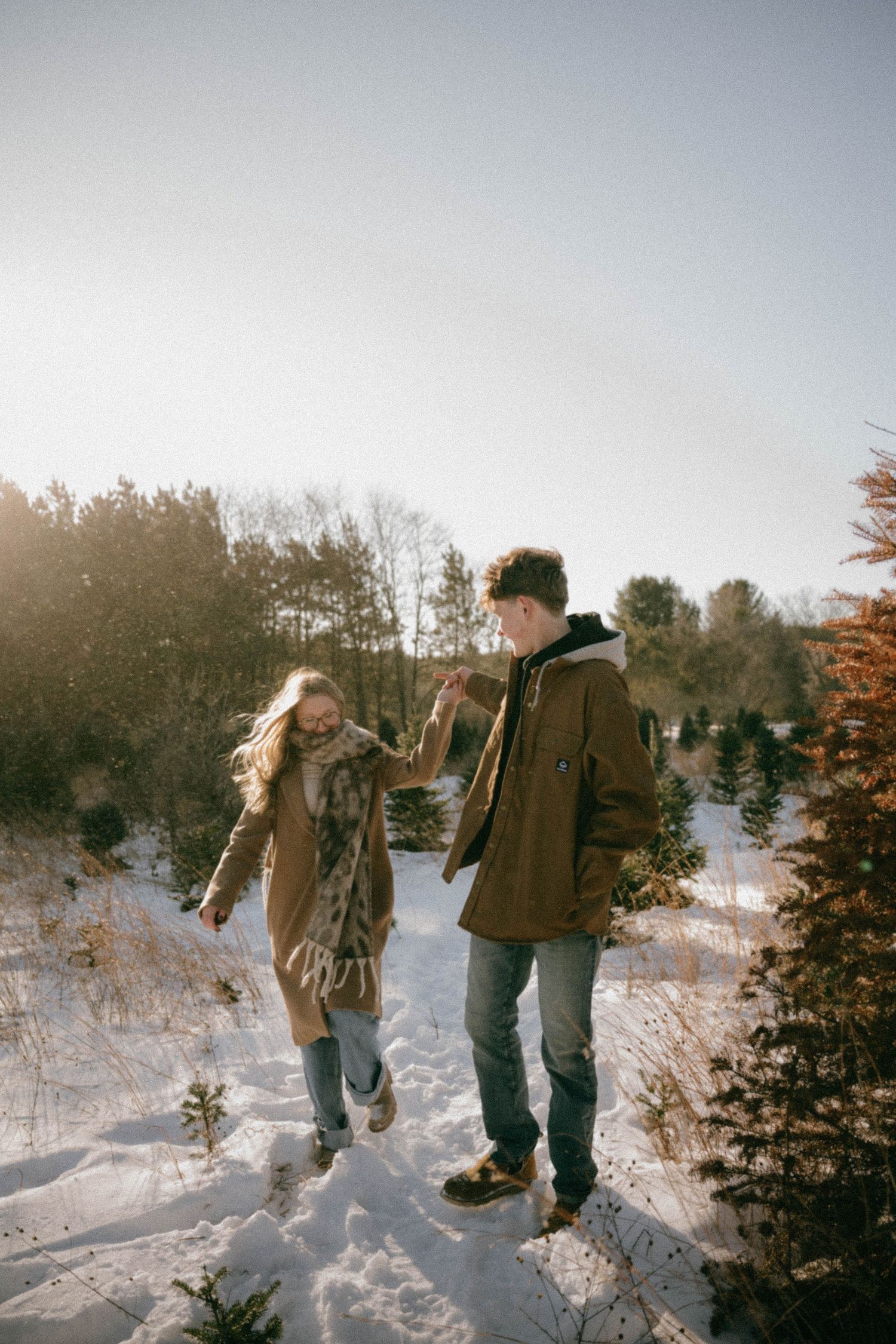 A happy couple holds hands and walks through a snowy Christmas tree farm in warm winter outfits.
