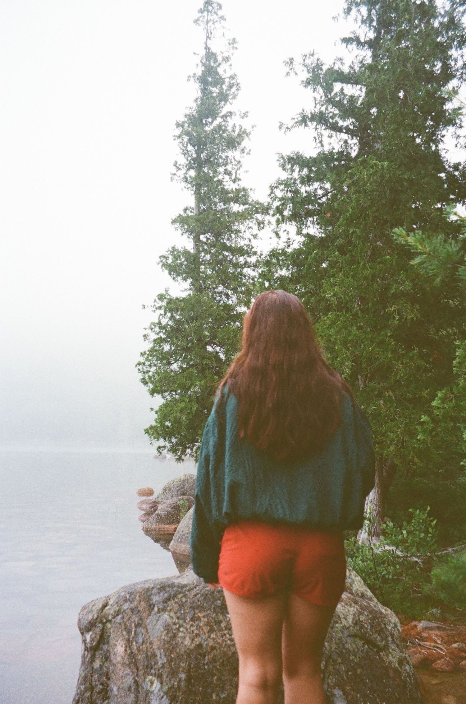 Woman with long hair in red shorts and green jacket standing on rocky misty lakeside overlook.
