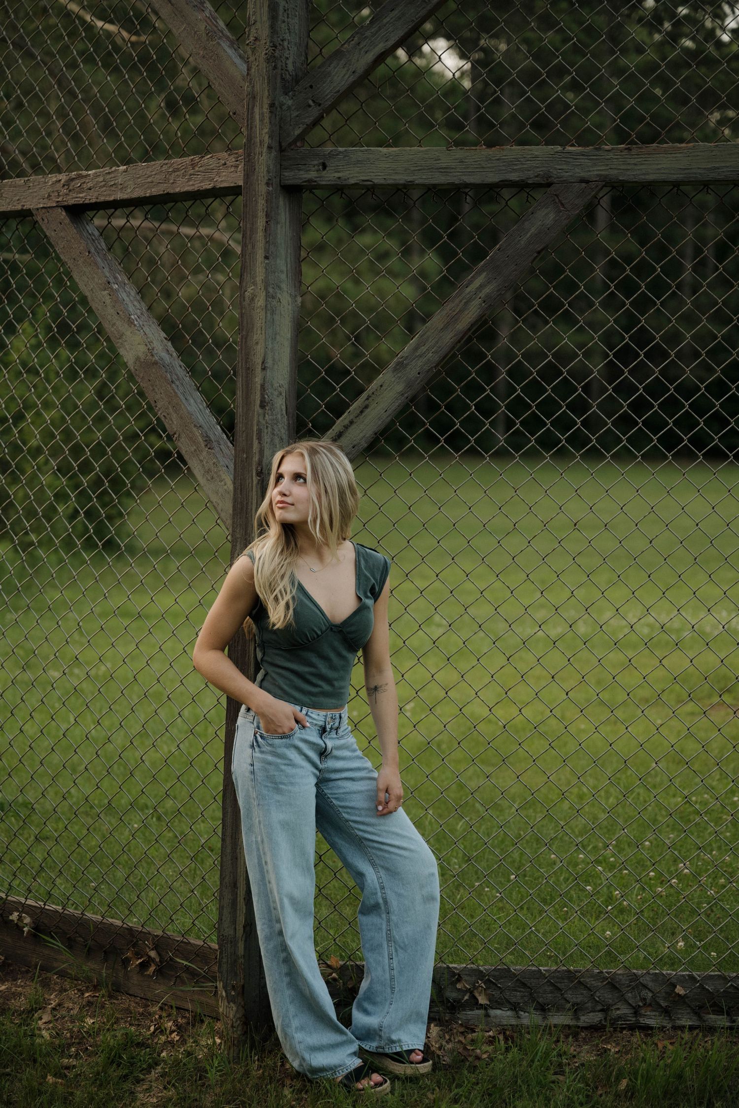 A blonde woman in jeans leans against a rustic chain-link fence in a green outdoor setting.