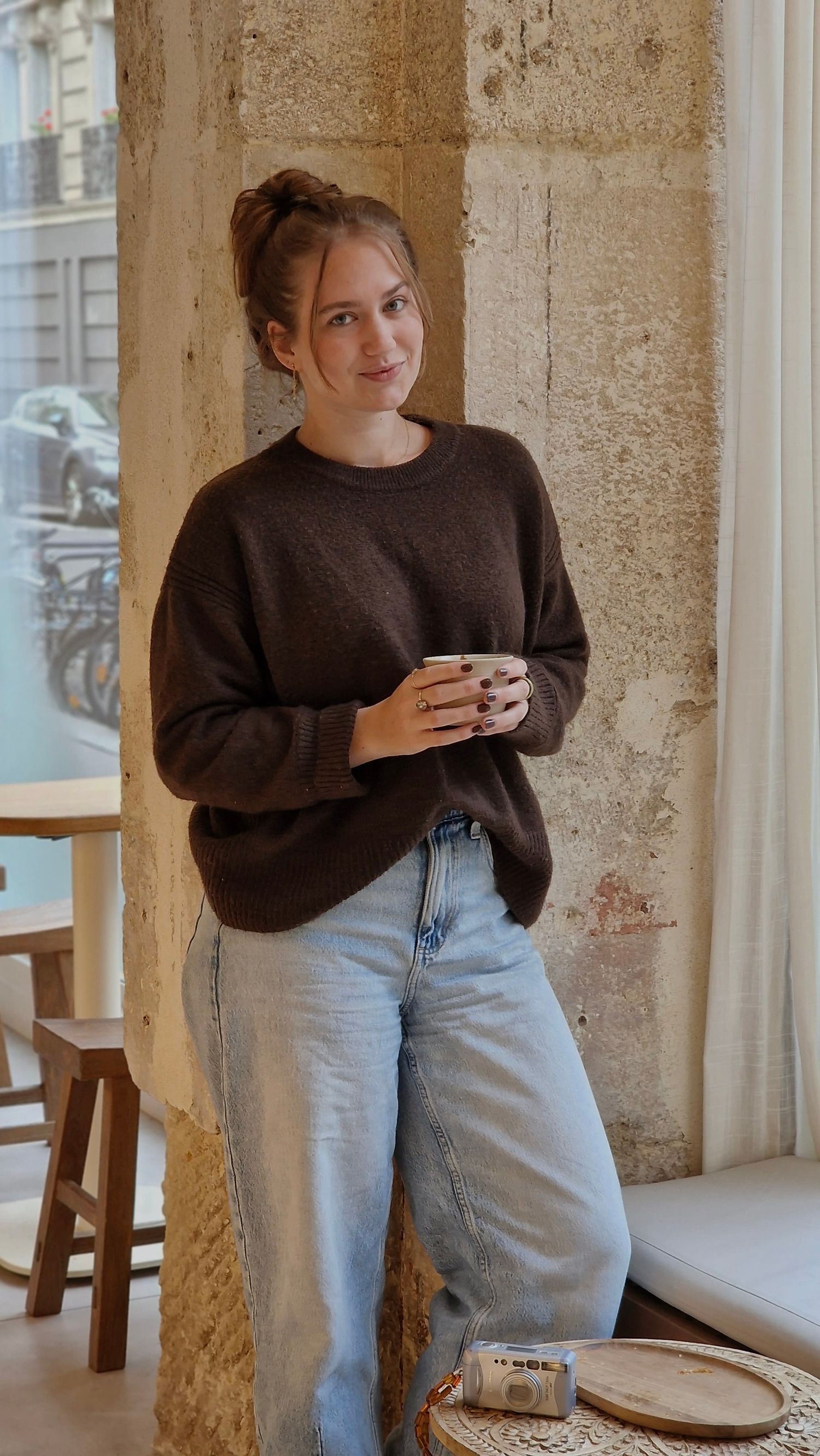 Young woman in brown sweater and jeans holding a coffee cup leaning against a stone wall in Paris cafe.