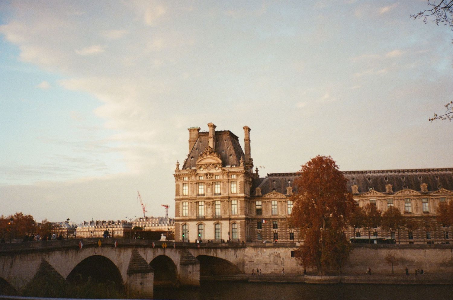 The Louvre museum building at golden hour reflected near a bridge over the Seine River in Paris.