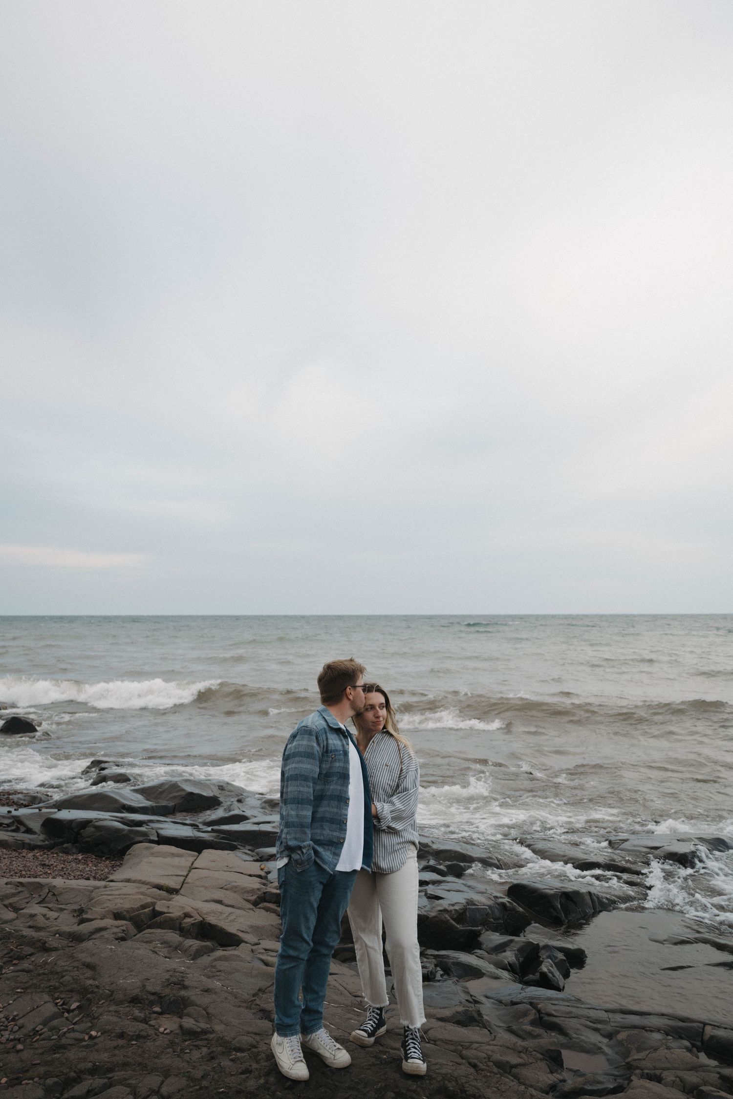 A couple embraces on rocky shoreline with waves crashing behind them under an overcast sky.