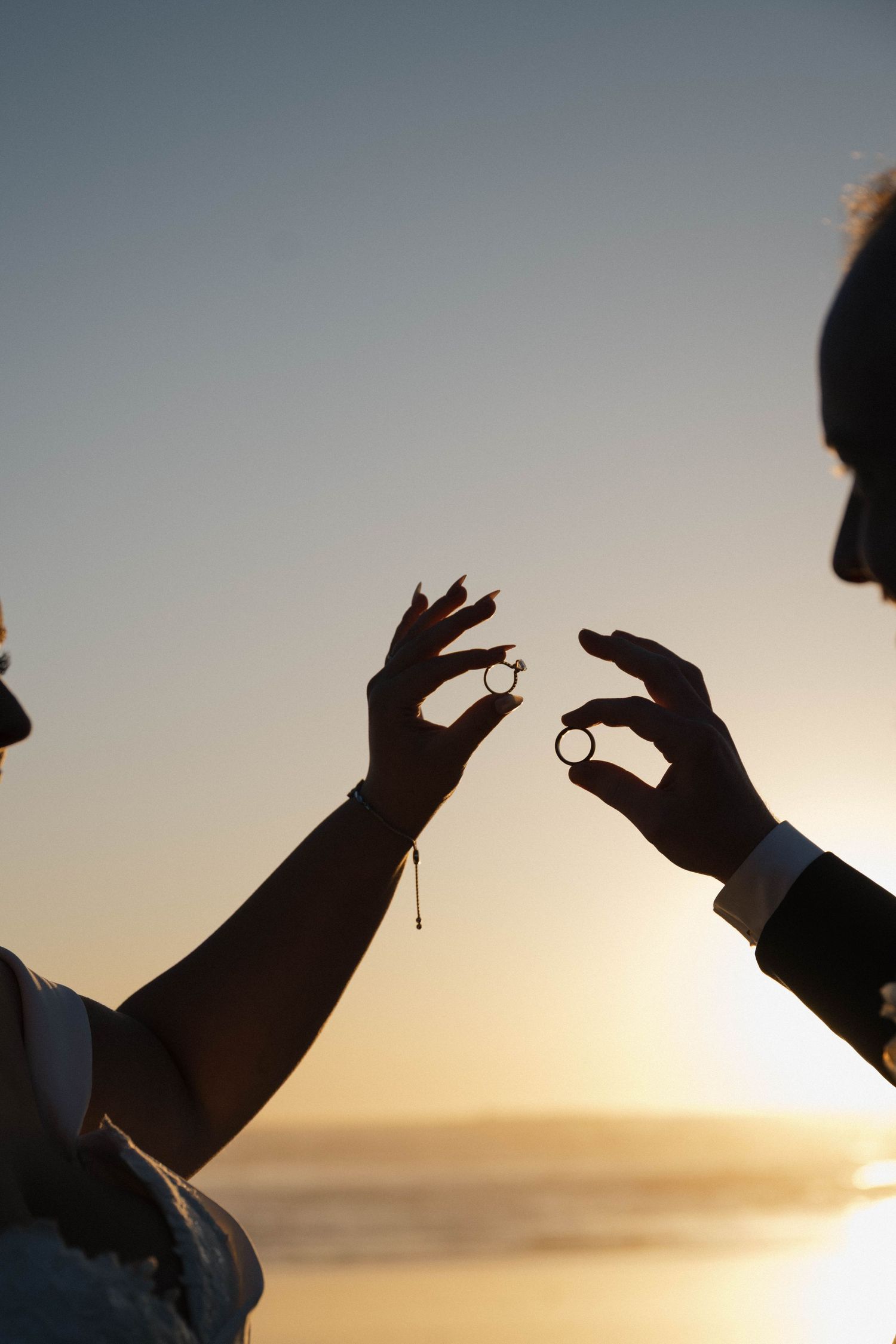 Silhouette of bride and groom holding wedding rings up against a glowing golden sunset over the ocean.