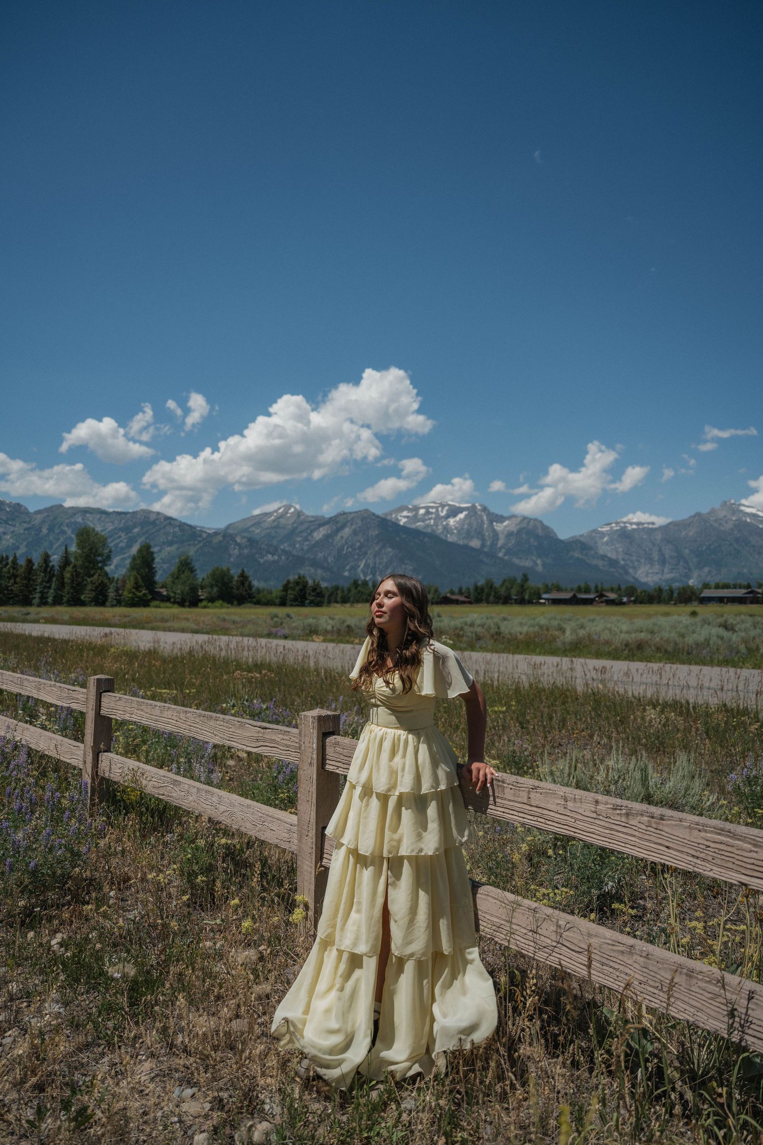 Woman in yellow tiered gown stands by rustic fence with Grand Teton mountains and blue sky backdrop.