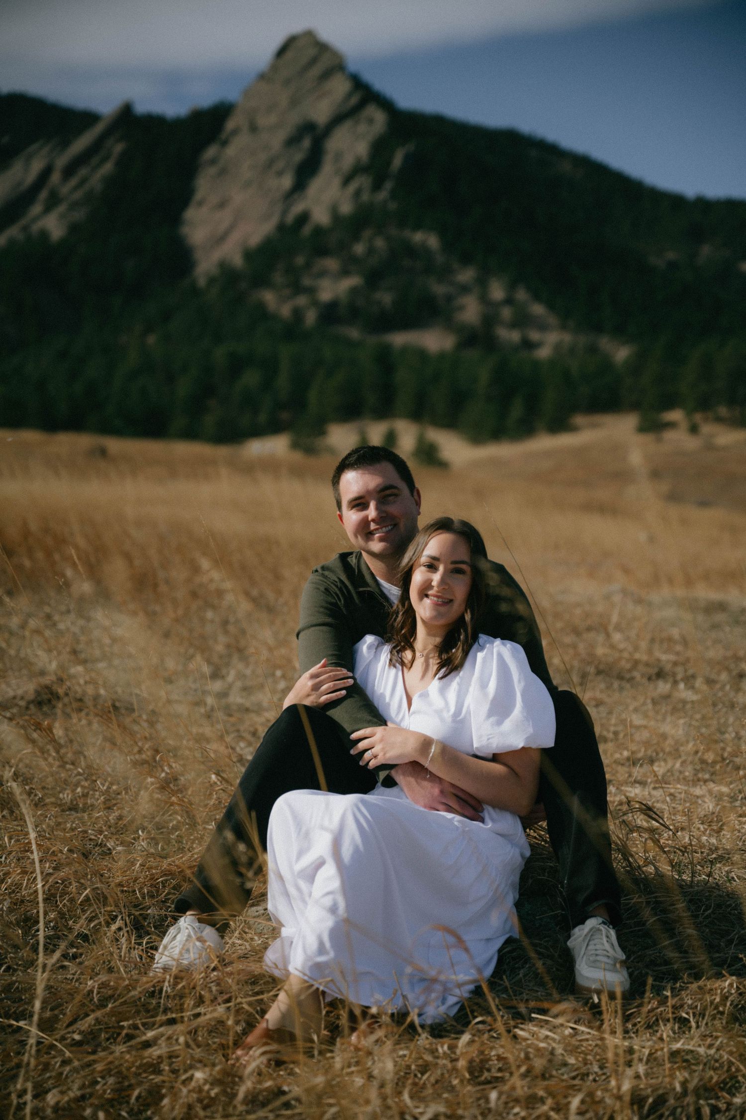 Smiling couple sits together in golden grass field with Rocky Mountain Flatirons behind them.