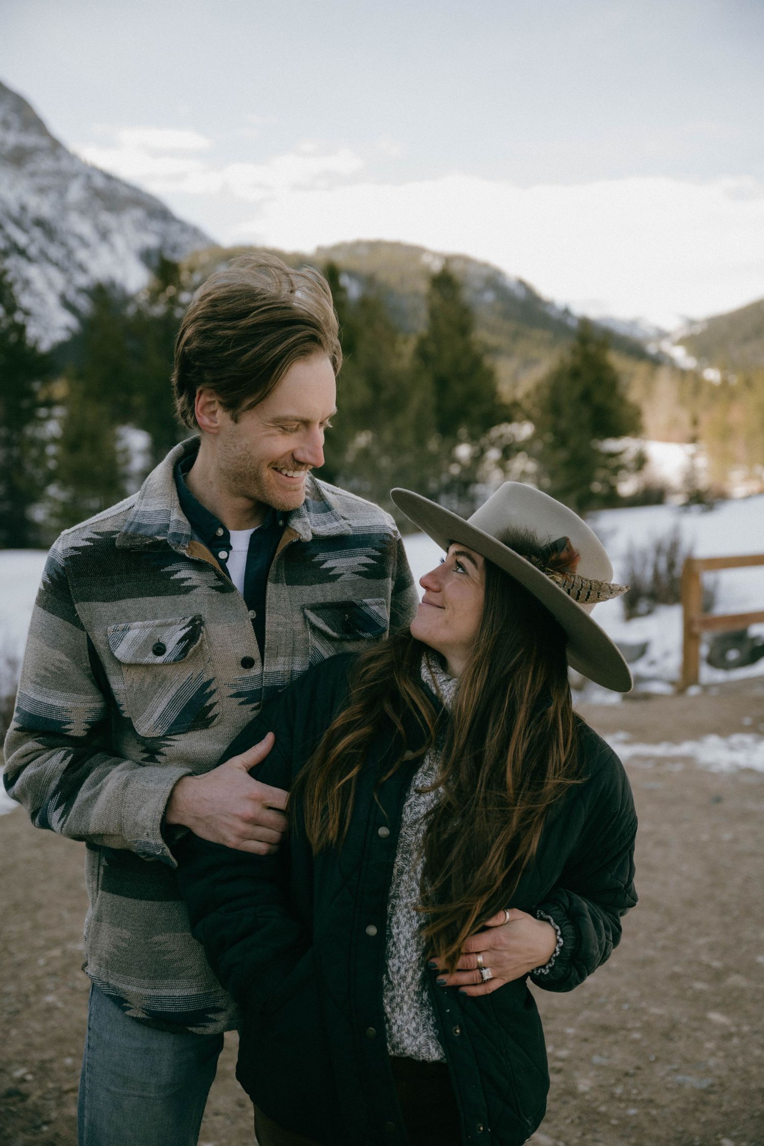 Smiling couple in winter attire with snowy mountains, woman wearing a wide-brim hat, looking at each other.