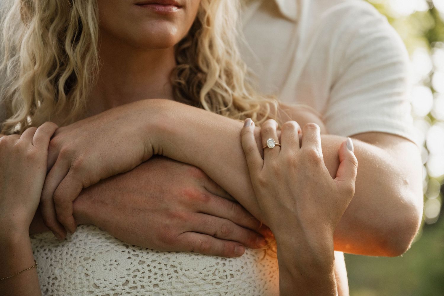 Close-up of couple embracing, woman wearing diamond engagement ring with man's arms wrapped around her.