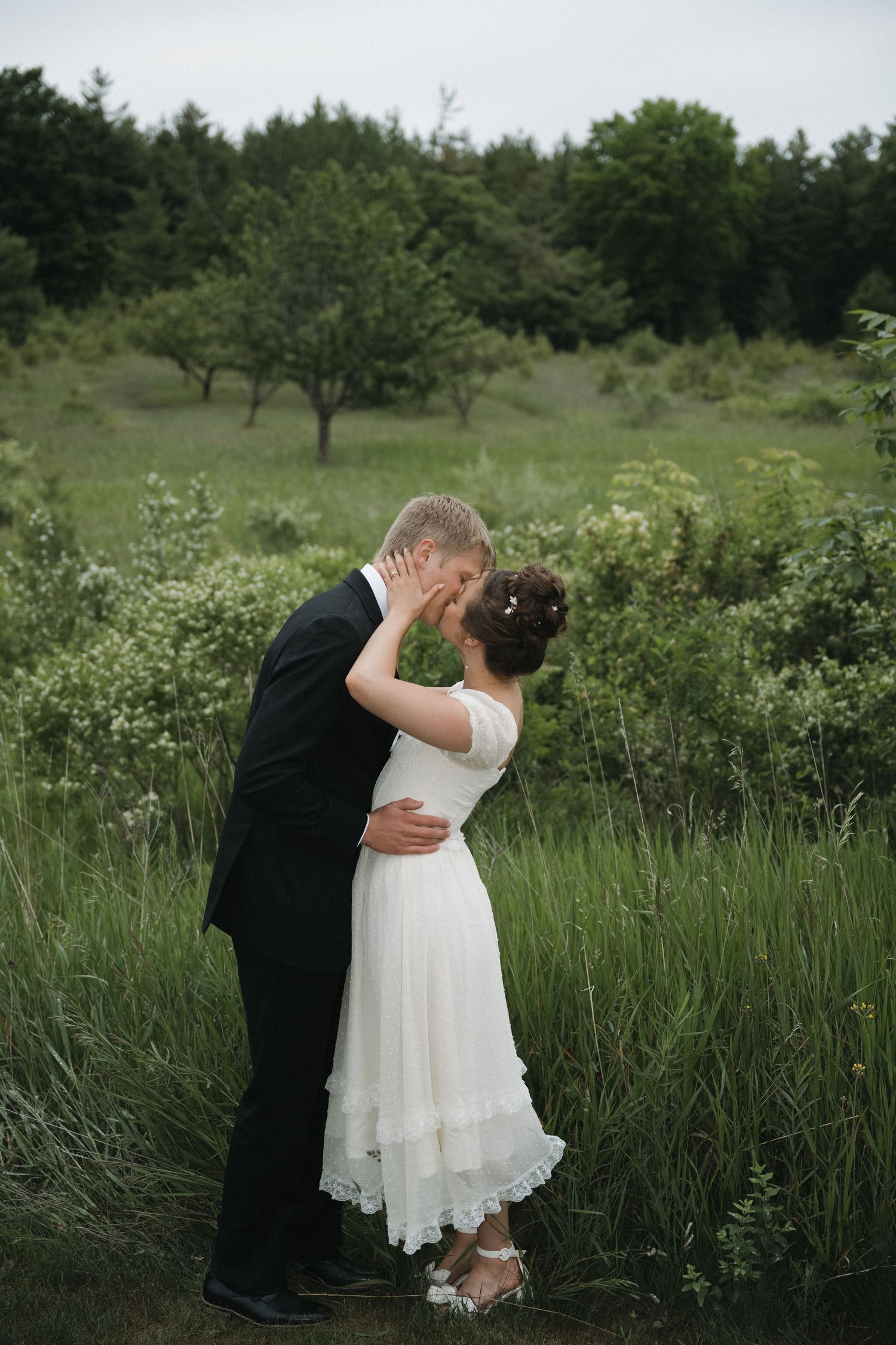 Wedding couple sharing a tender kiss outdoors in a green field, bride in white lace dress and groom in classic black suit.