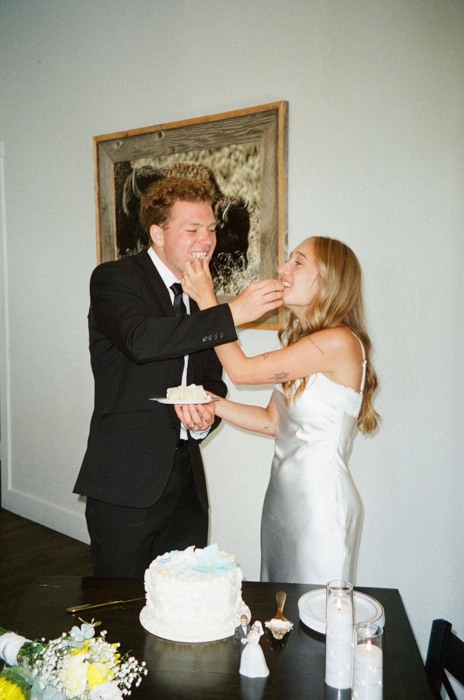 Bride and groom playfully feed each other wedding cake at indoor reception celebration.