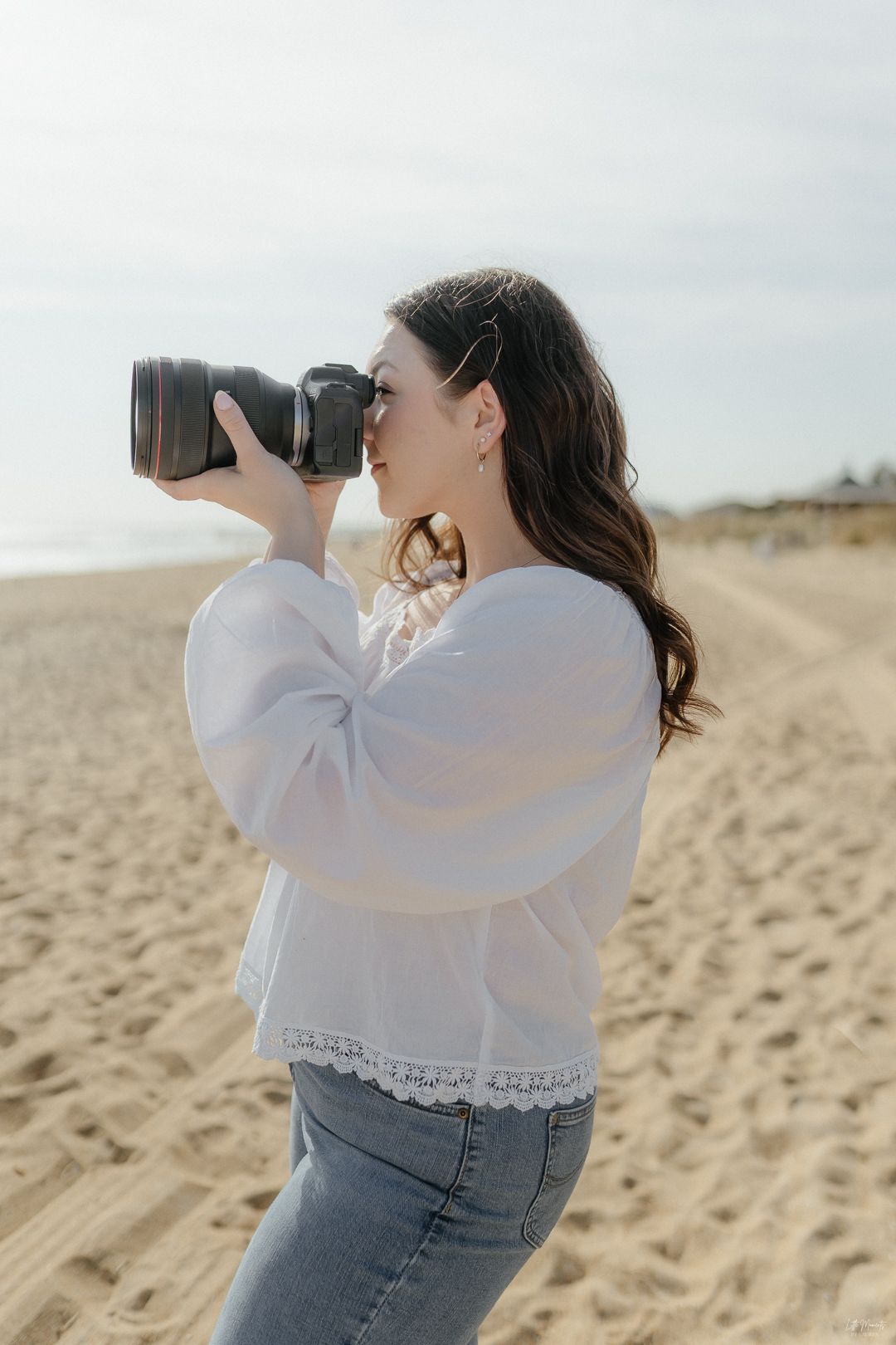 Female photographer taking photos with Canon camera on sandy beach in white blouse.