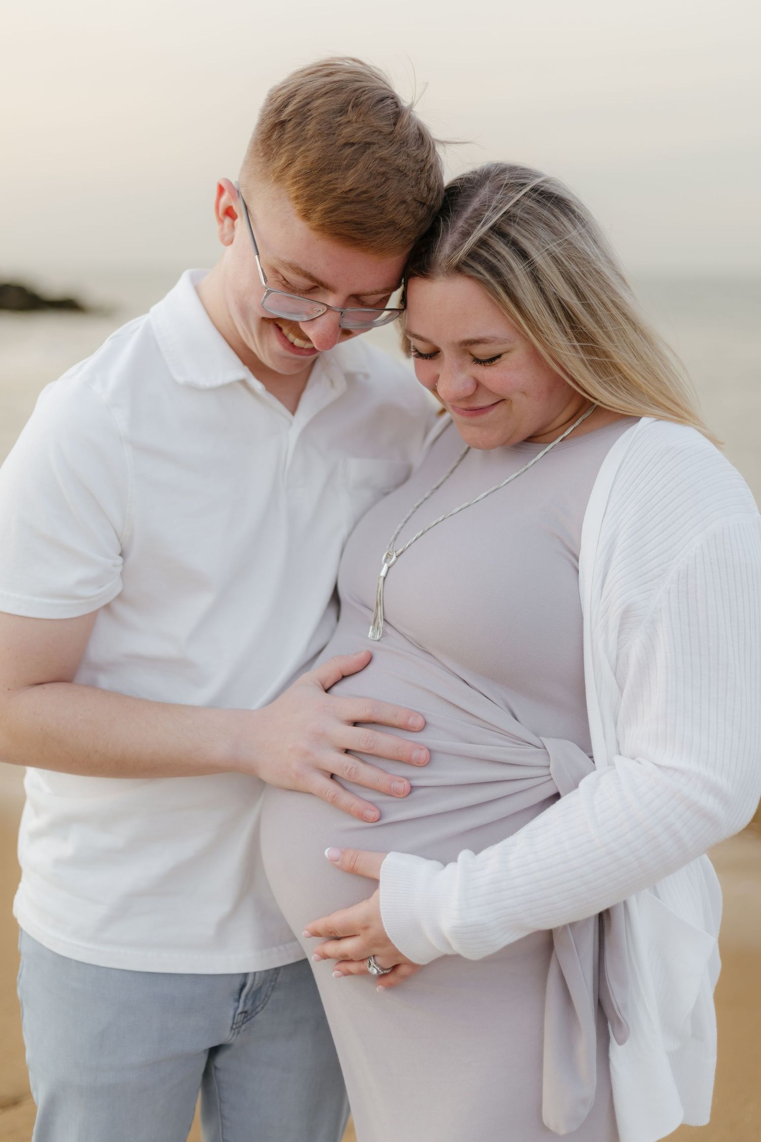 Smiling couple cradling pregnant belly together on sandy beach at sunset.