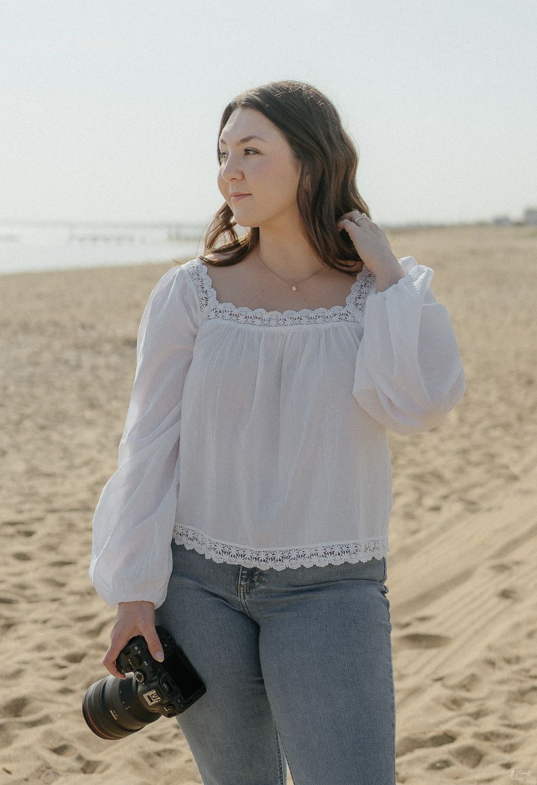 Brunette female photographer standing on beach holding Canon camera in white top and jeans.