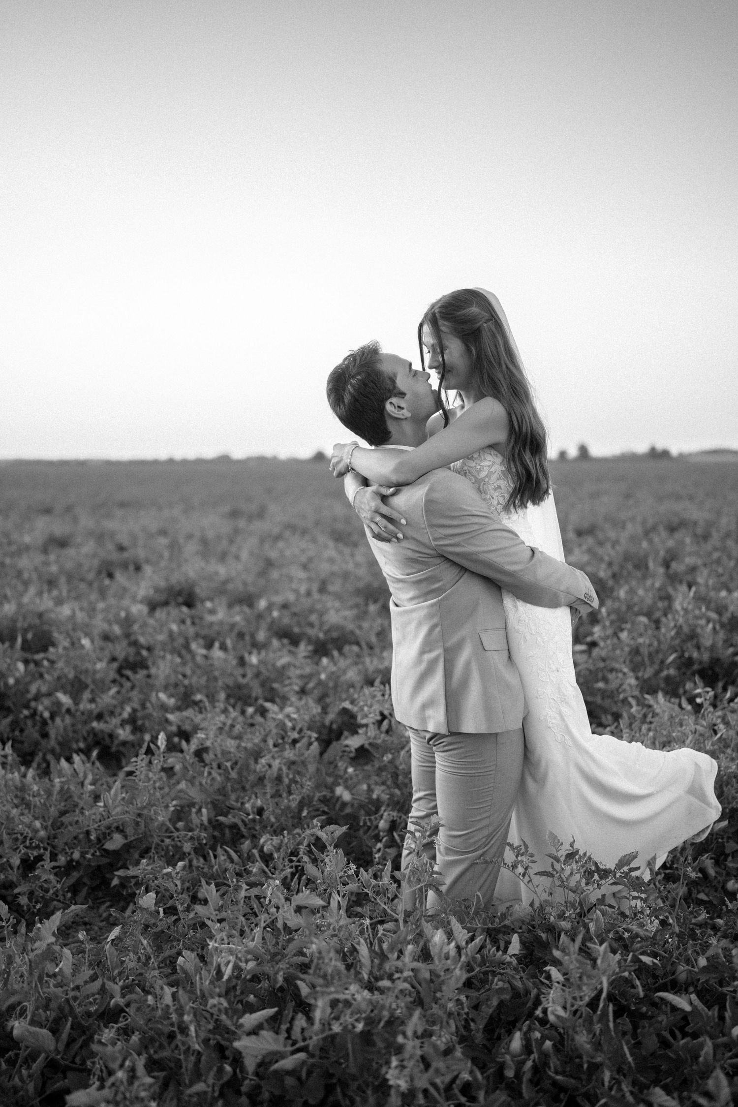 Black and white photo of couple embracing in flowering field during sunset.