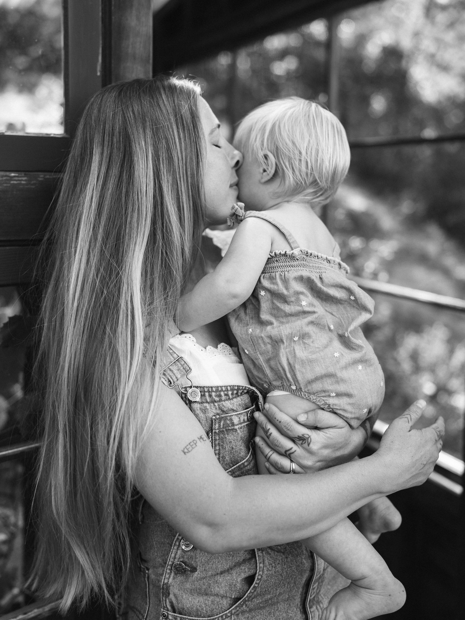 Black and white photo of a mother with long hair kissing her toddler child while holding them outdoors.