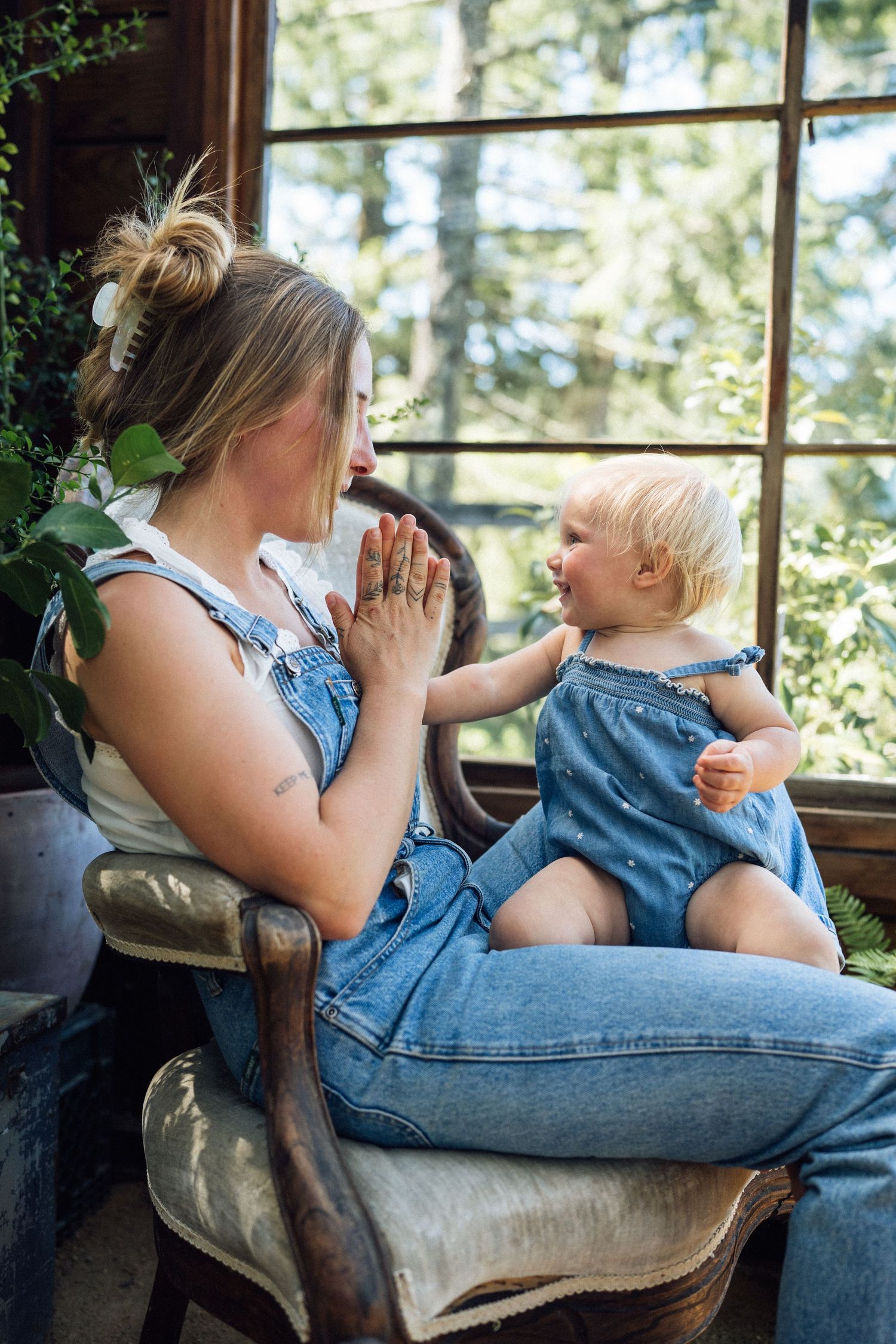 Mother and baby girl in matching denim overalls sitting by a sunlit window, sharing a playful moment together.