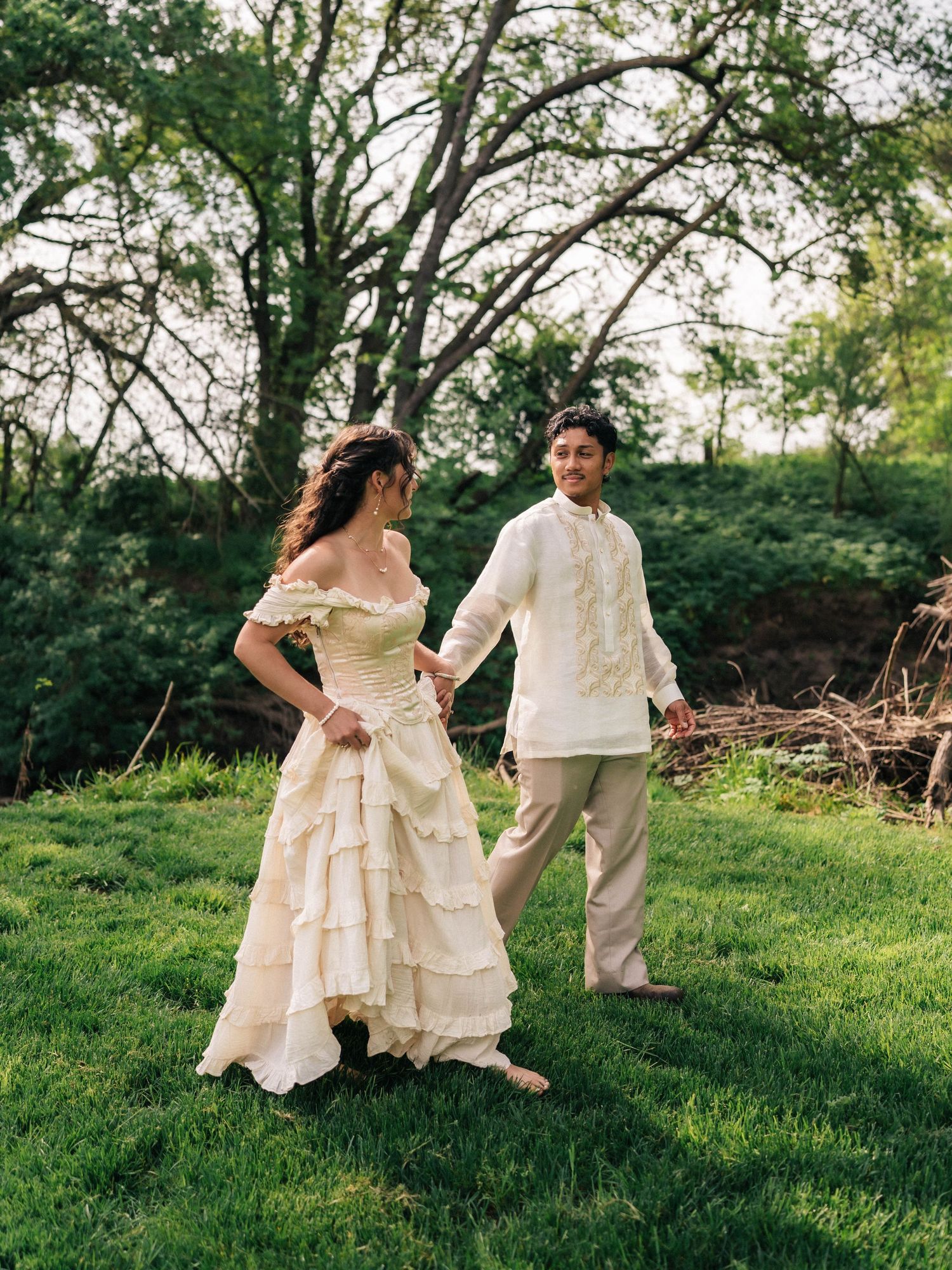 Couple in traditional Filipino wedding attire dancing on green grass with lush trees in the background.