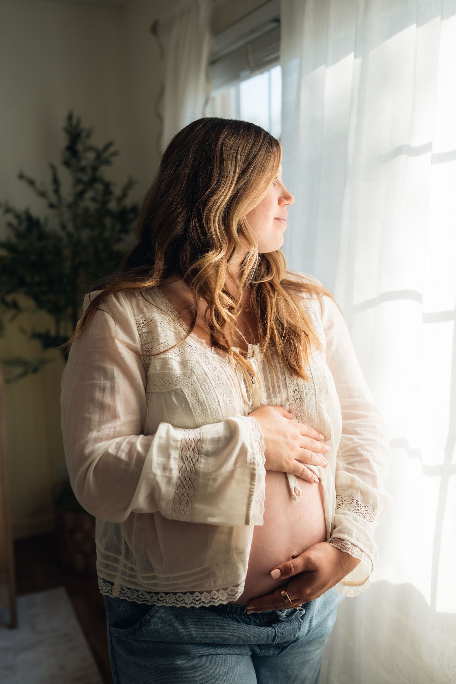 Pregnant woman in white lace blouse and jeans cradling her baby bump while gazing out a sunlit window.