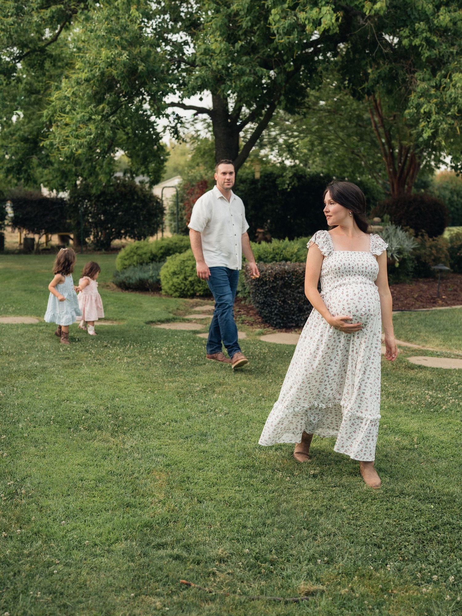 Pregnant woman in floral dress walks barefoot on grass with man in white shirt and two young girls in a lush garden.
