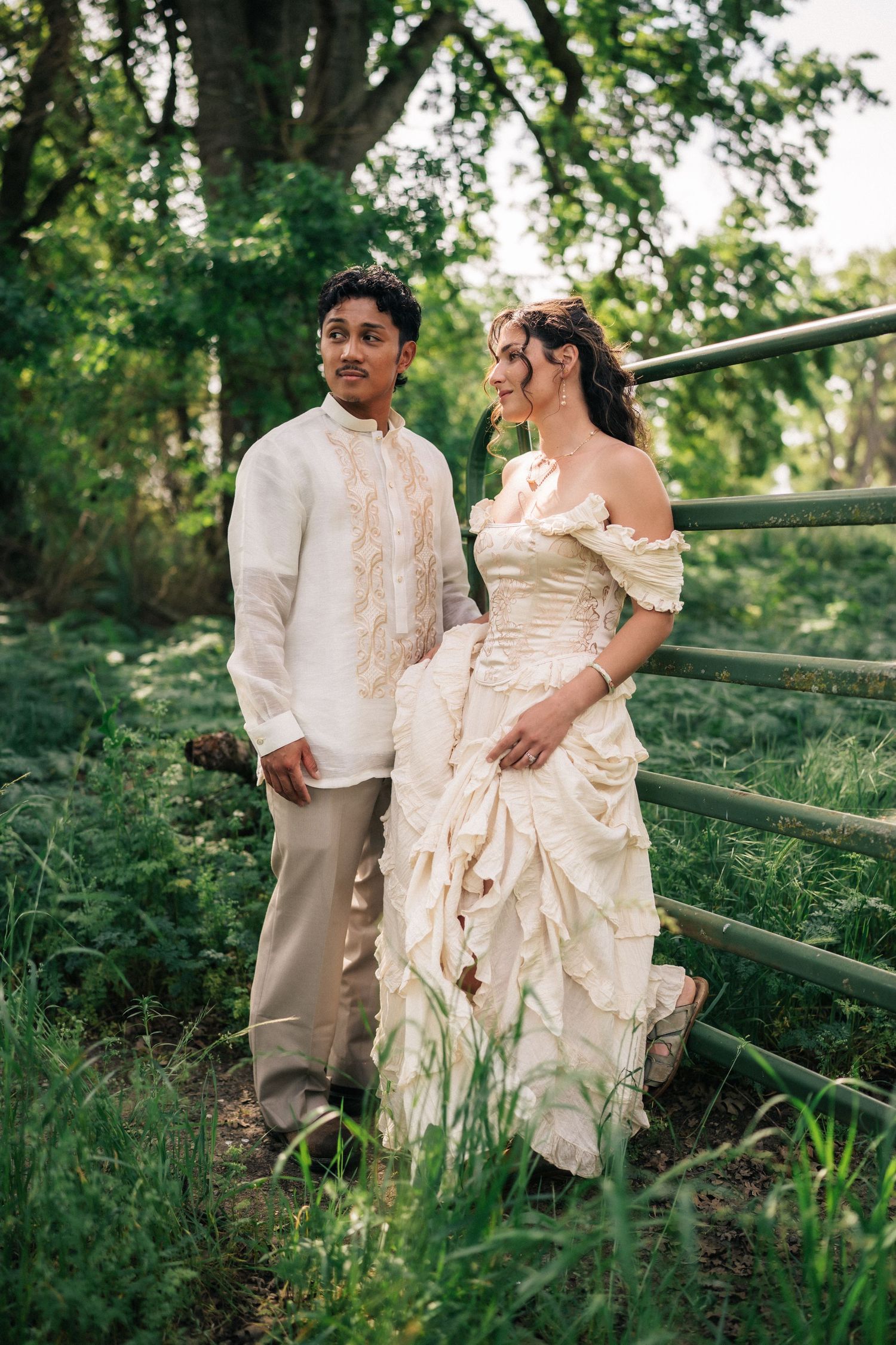 Couple in elegant cream wedding attire posing by a rustic fence surrounded by lush green trees outdoors.