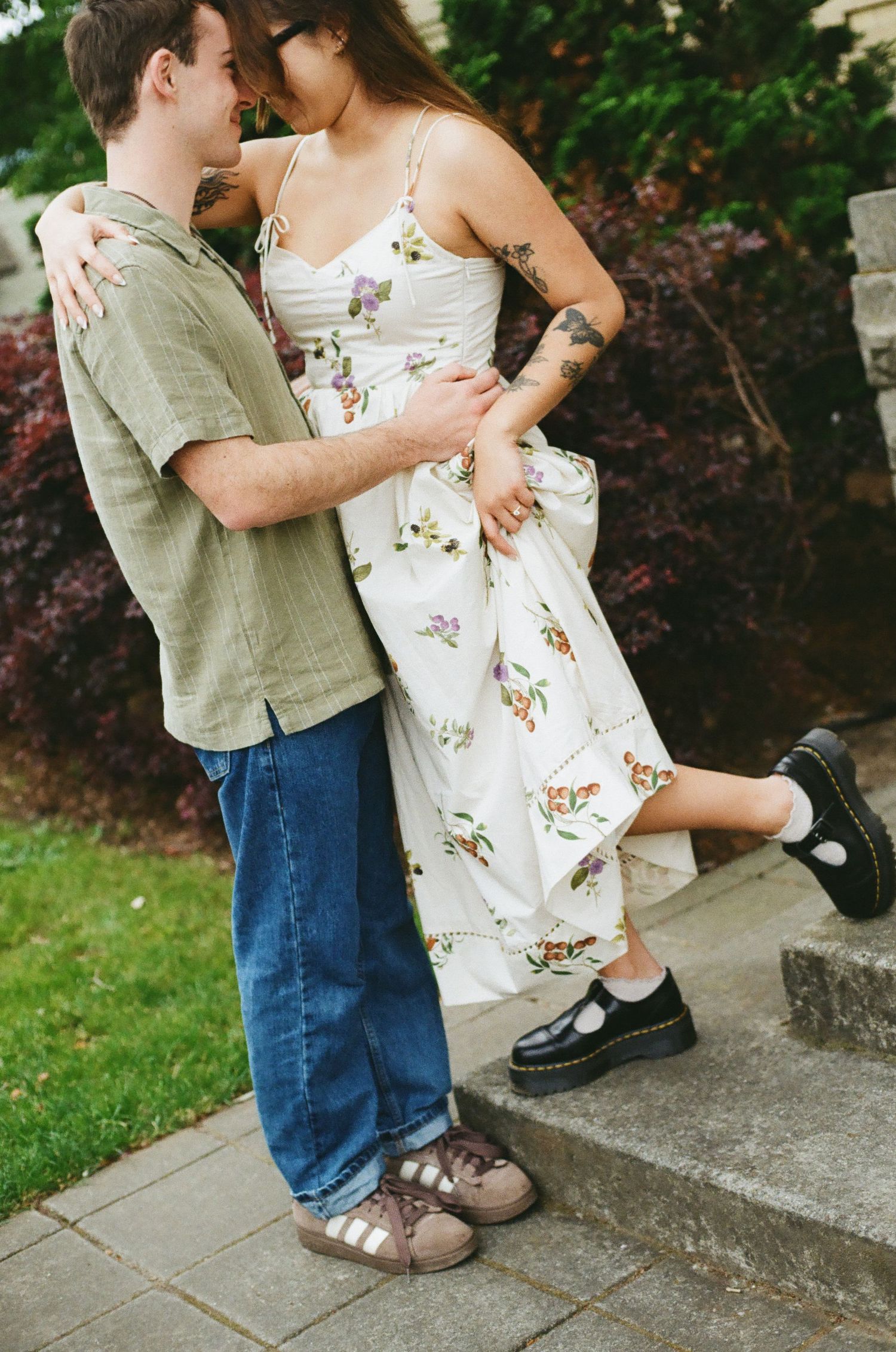 A couple embraces outdoors, woman in floral dress and Dr. Martens, man in green shirt and jeans, standing on stone steps.