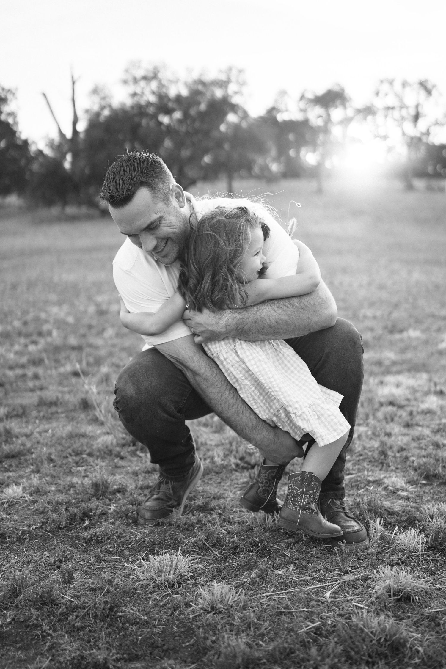 Black and white photo of a father crouching to hug his young daughter in a sunlit field.