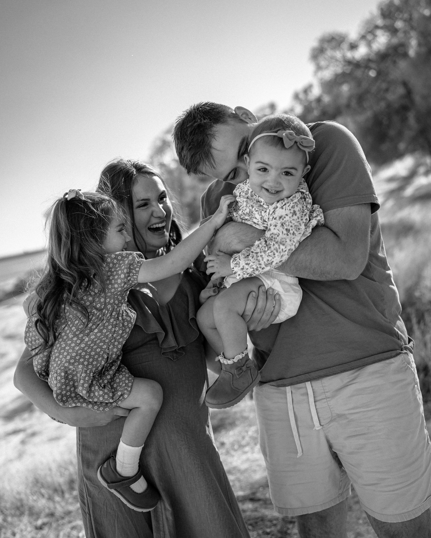 Black and white family photo of parents laughing and holding two young daughters outdoors in a sunny field.