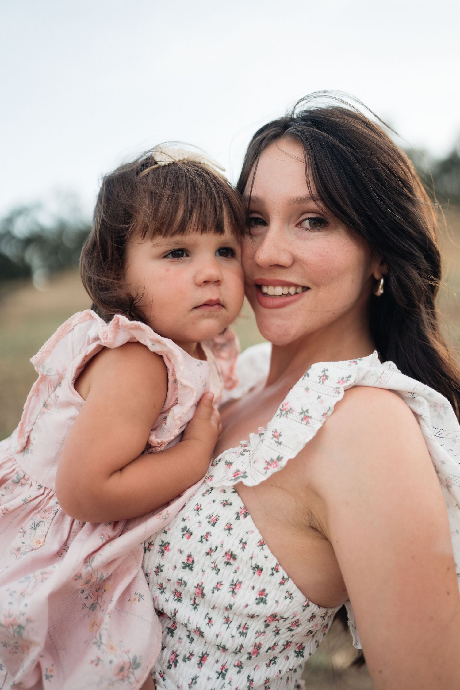 Young woman in floral dress smiling while holding a toddler girl in pink dress outdoors.