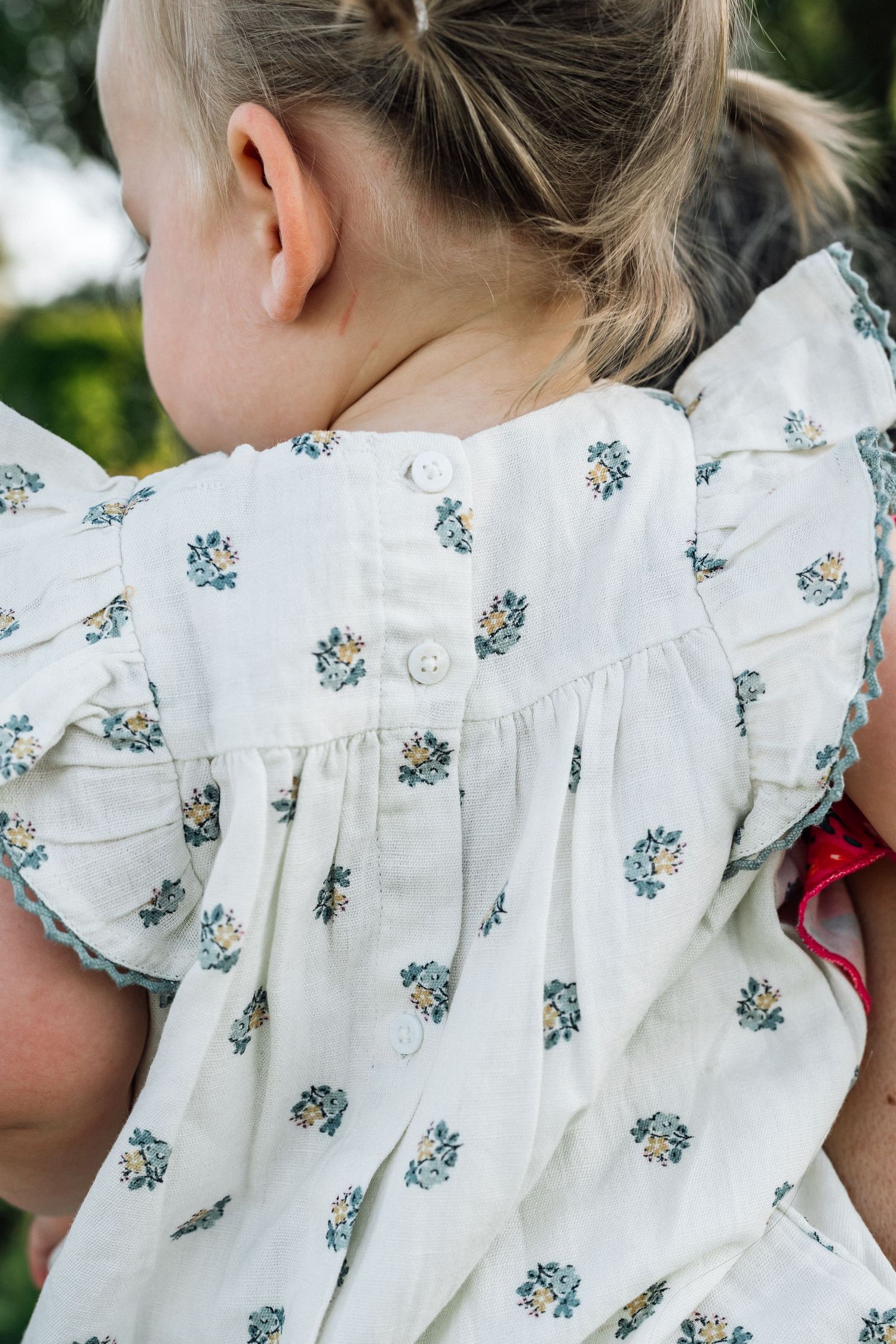 Young girl wearing a white floral ruffle sleeve dress with button back detail and blue trim accents.