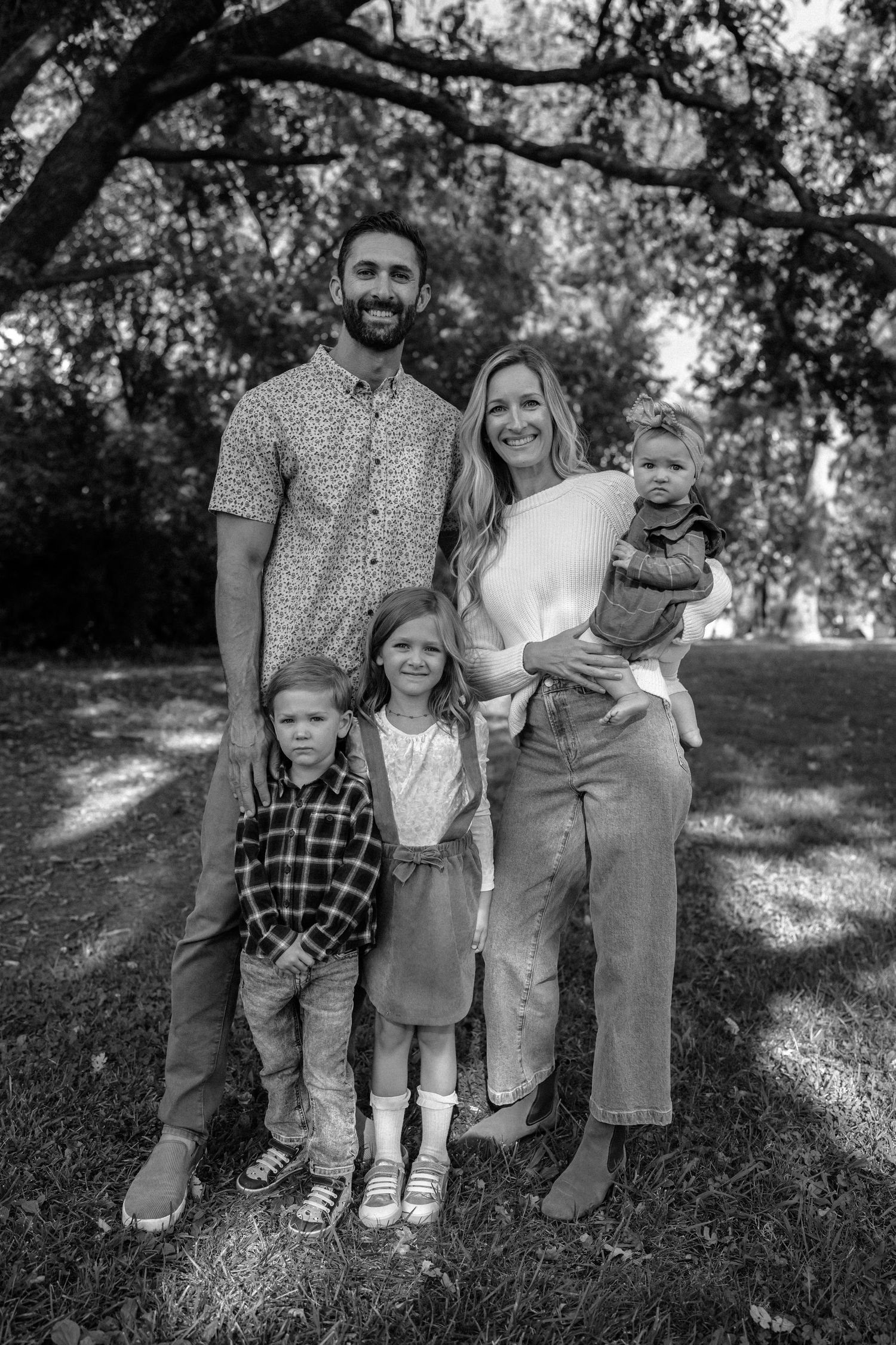 A family portrait captured under a tree with dappled sunlight in black and white.