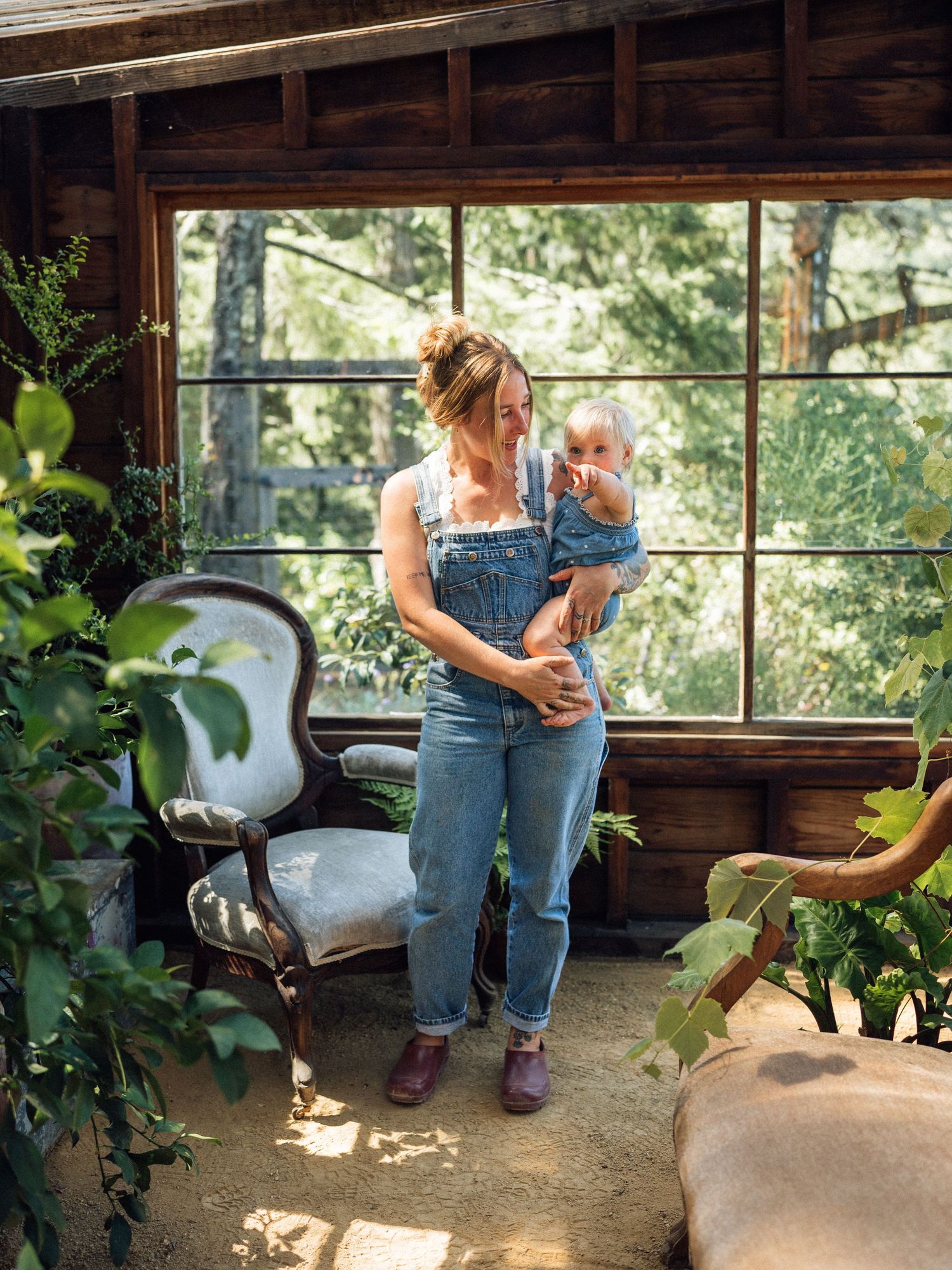 Woman in denim overalls holding a baby in a rustic sunroom surrounded by lush plants and vintage furniture.