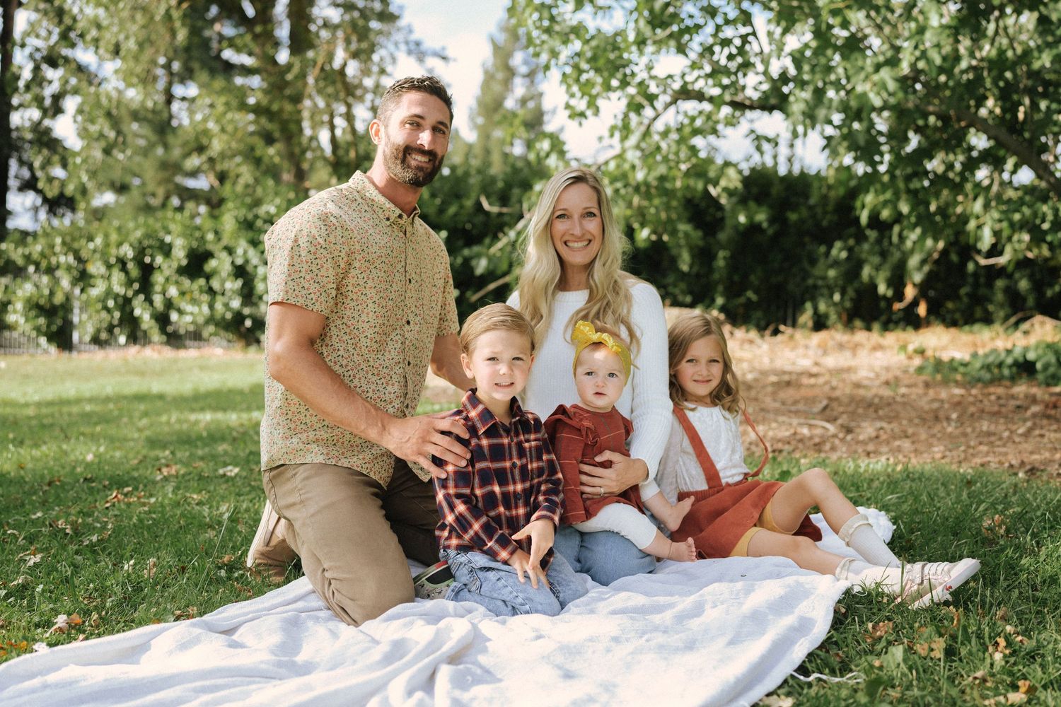 Family enjoying a picnic together on a blanket spread out on green grass under trees on a sunny day.