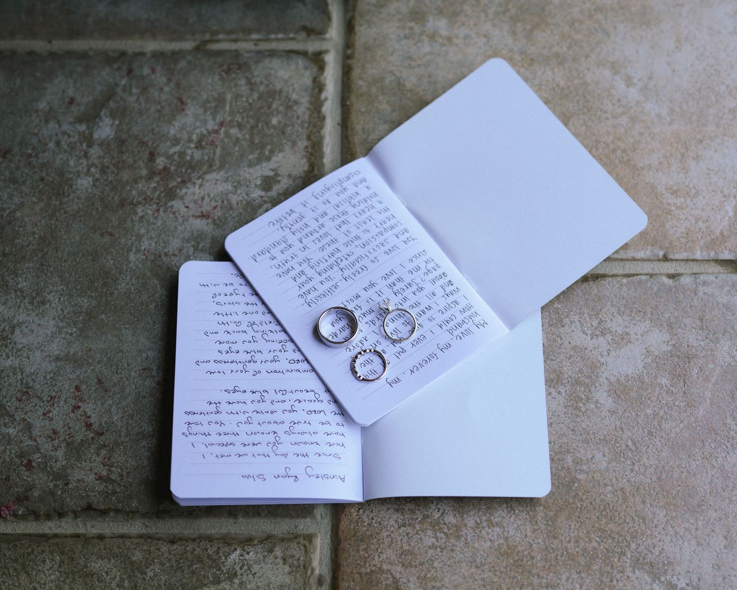 Wedding rings rest on handwritten vows in a white notebook against a stone tile background.