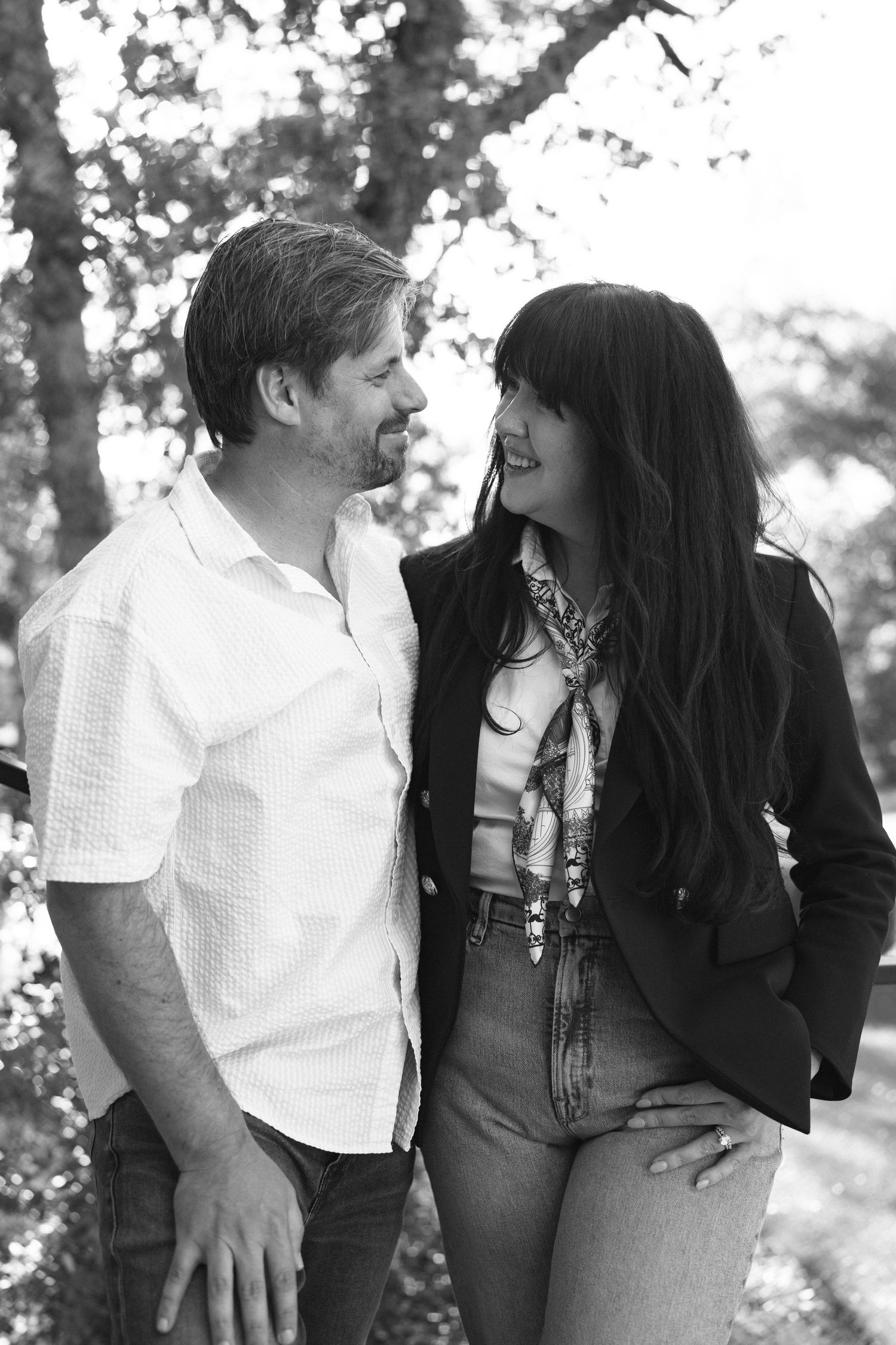 Black and white photo of a smiling couple standing together outdoors among trees, woman with dark hair wearing blazer and scarf.