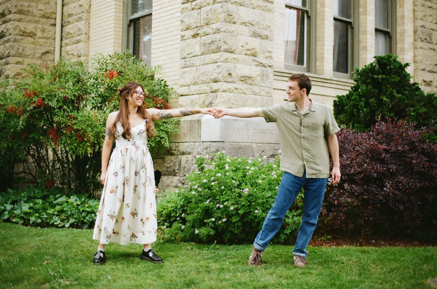 Couple holding hands and dancing outdoors in front of a stone building surrounded by lush green gardens.