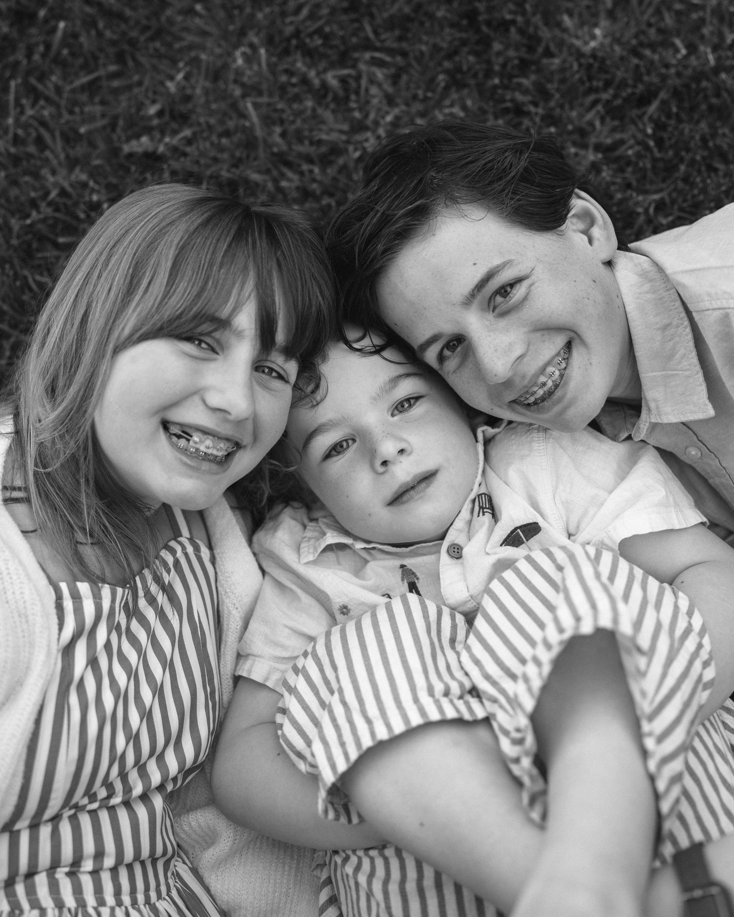 Black and white photo of three smiling children lying on grass, two with braces, wearing striped shirts.