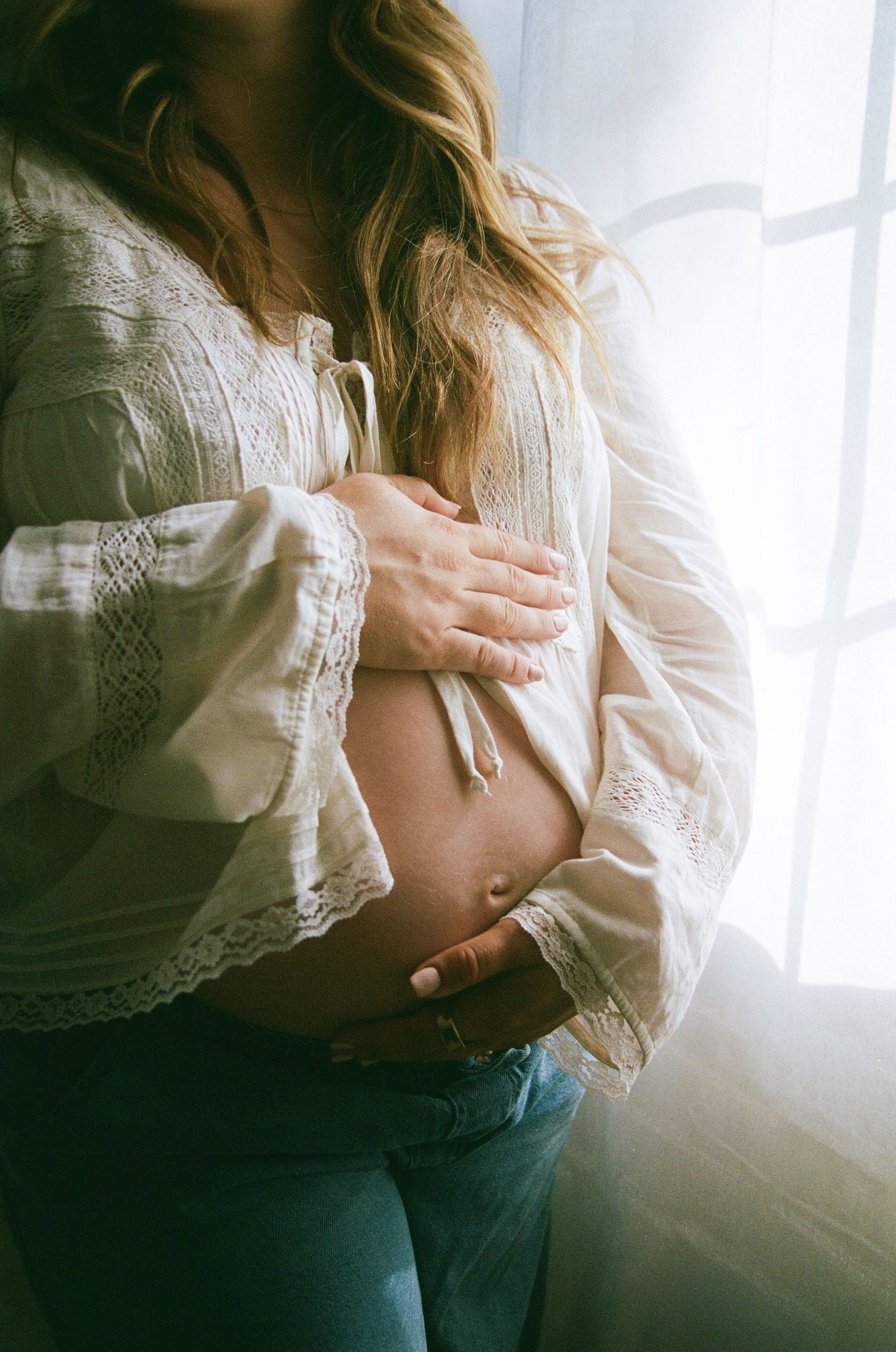 Pregnant woman in white lace blouse and jeans cradling her baby bump near a sunlit window.