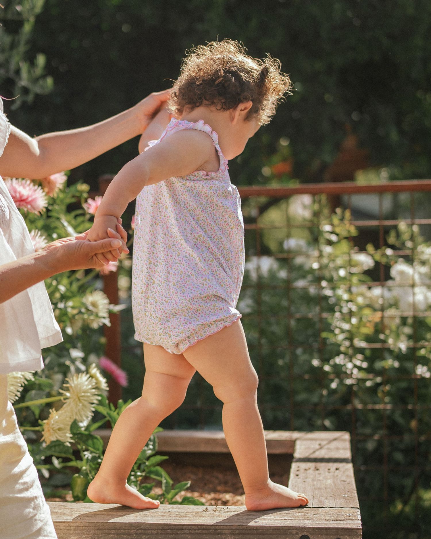 Small bare feet walking on wooden steps in a garden setting with lush greenery and flowers in the background.