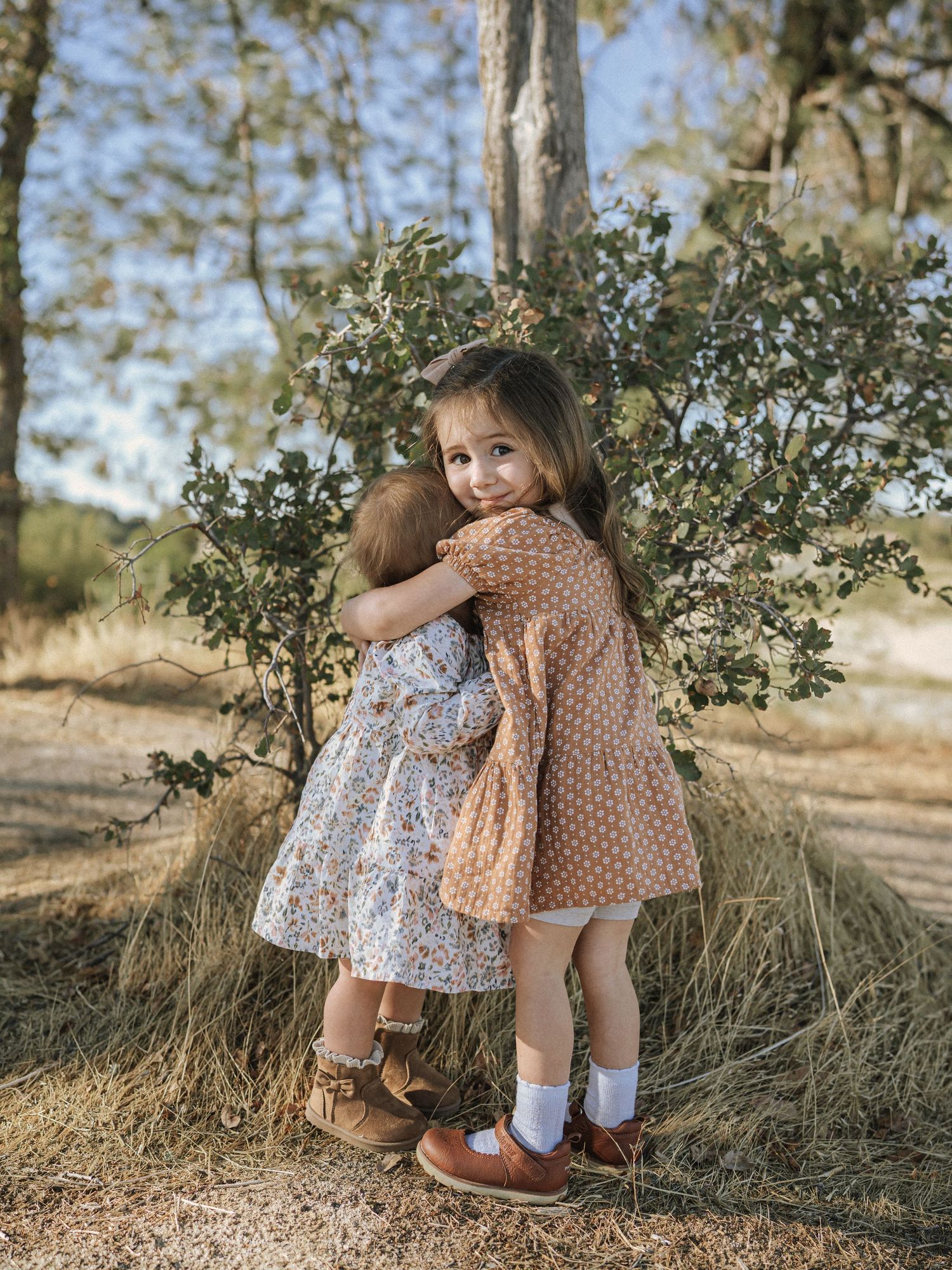 Two children in vintage-style dresses playing together near bushes in natural outdoor setting.