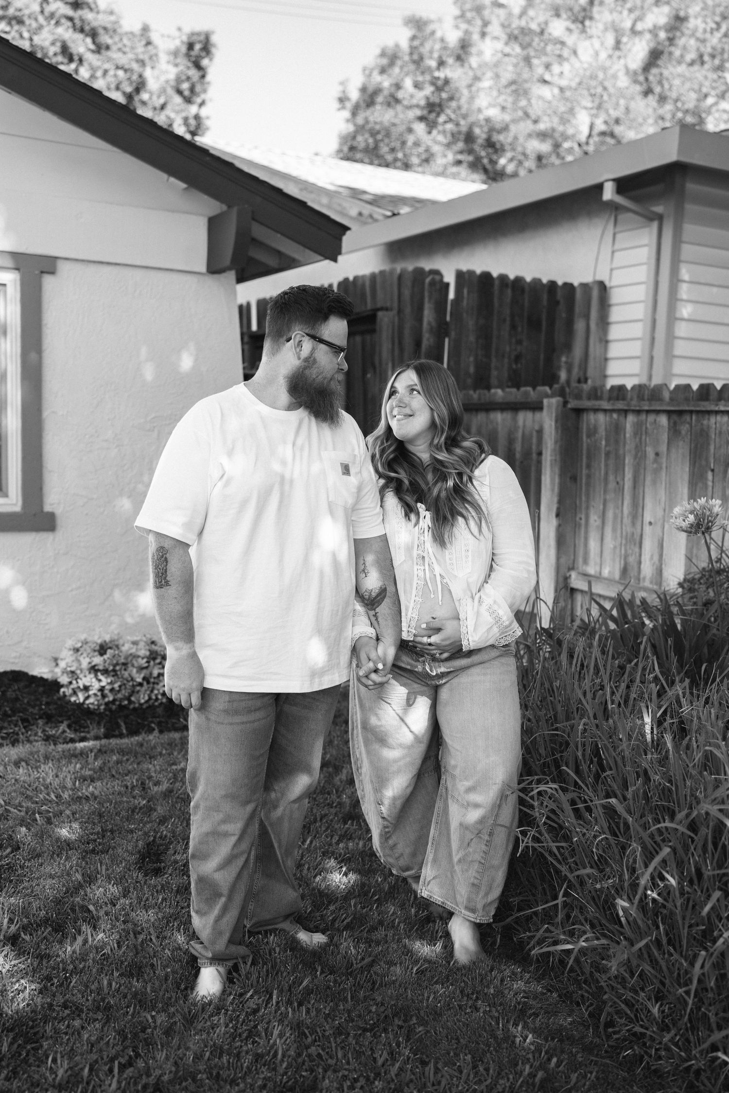Black and white photo of a smiling couple holding hands in a backyard, wearing casual clothes, standing near a house.