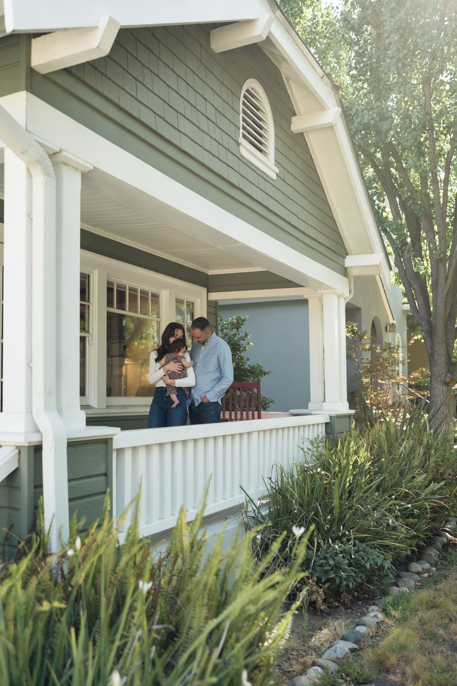 Young family with baby standing on front porch of charming green craftsman style home surrounded by lush greenery.