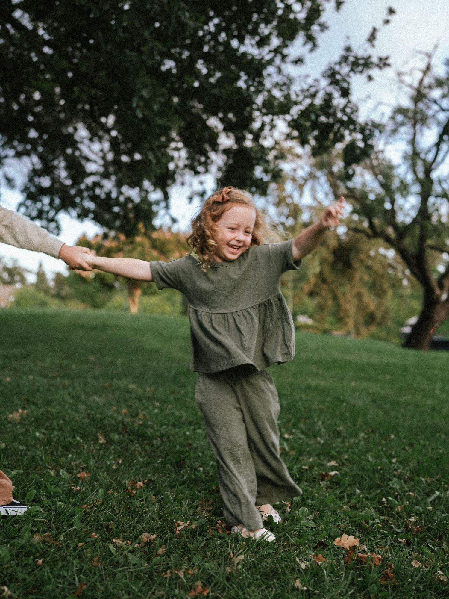 Child in olive green outfit playing and twirling joyfully on grassy hillside.