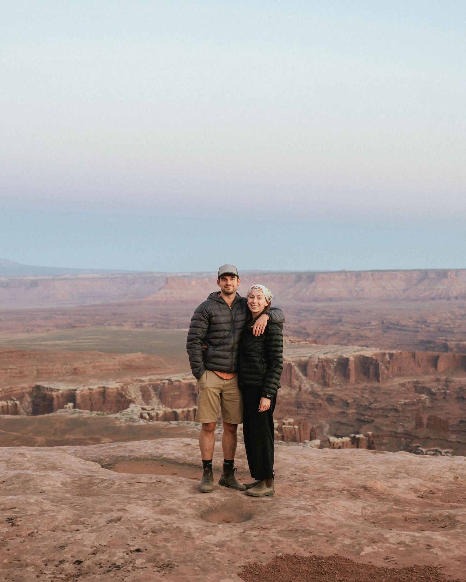 Two people pose together on a cliff edge overlooking a vast desert canyon landscape.