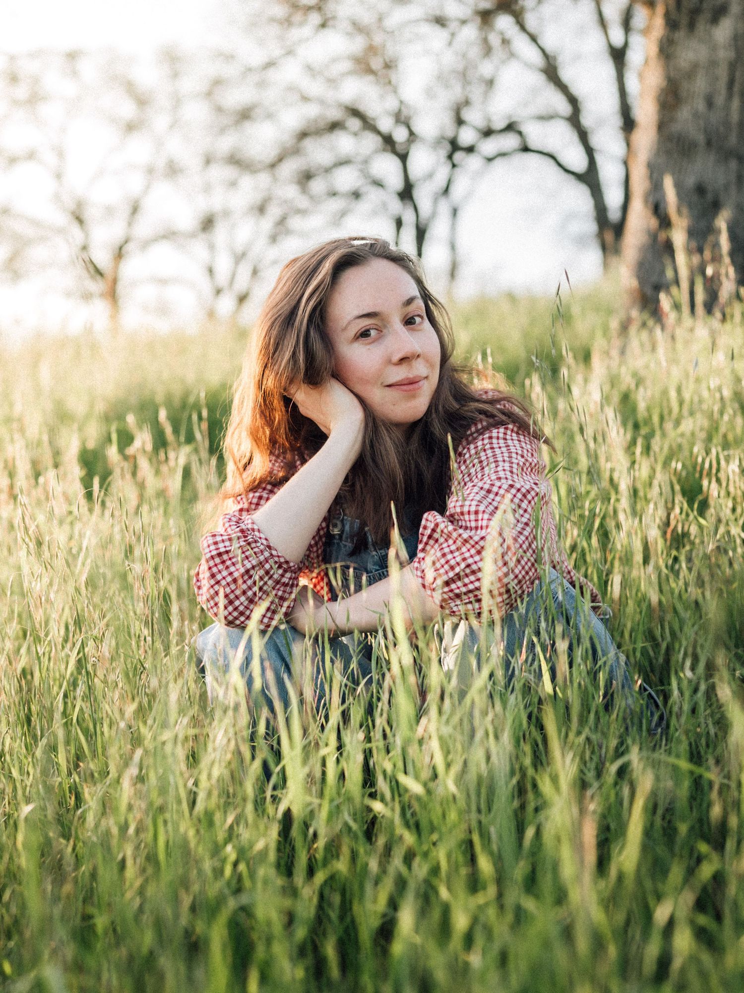 Person in red plaid shirt sits thoughtfully in tall grass field during golden hour with trees in background.