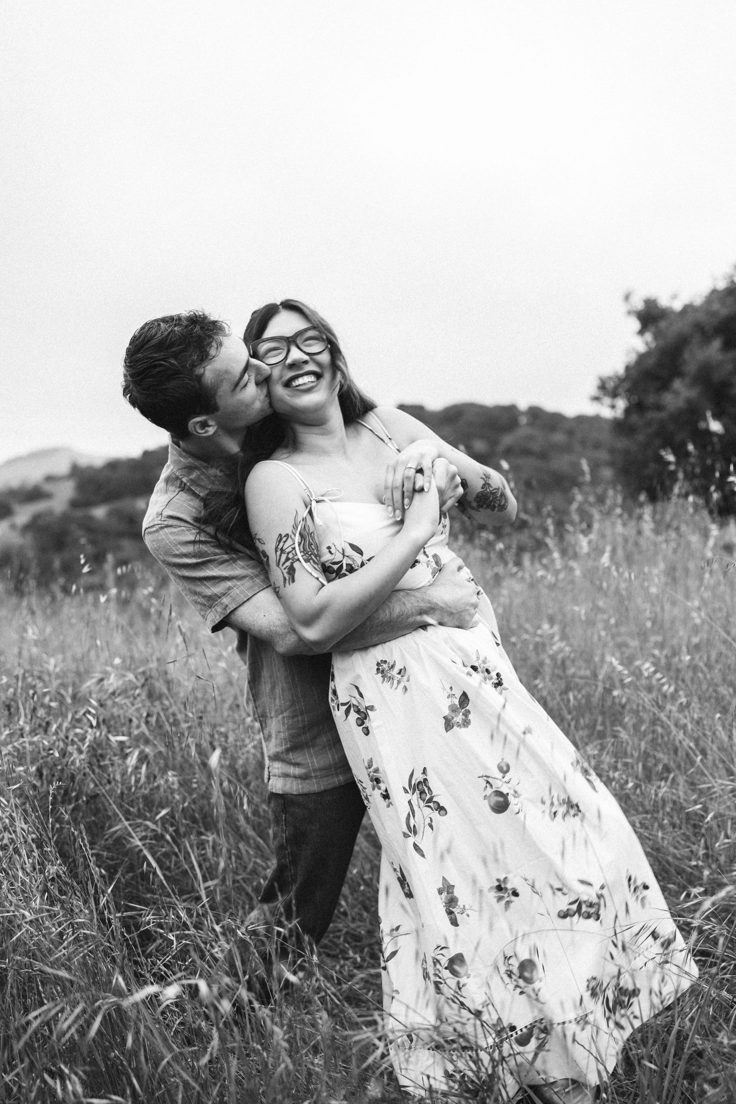 Black and white photo of a couple embracing in a field, man kissing woman's cheek as she laughs joyfully.